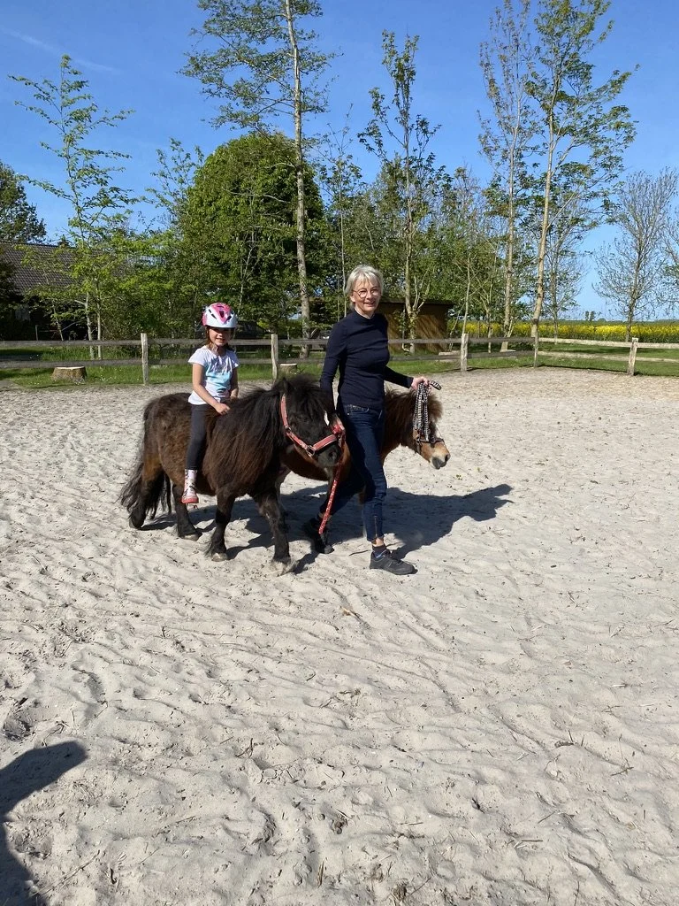 Ein junges Mädchen mit Helm sitzt auf einem Pony, während eine ältere Frau neben einem zweiten Pony steht, beide Ponys sind auf einem Reitplatz mit Sandboden. Im Hintergrund sind Bäume und ein Zaun, der Reitplatz liegt unter einem blauen Himmel.