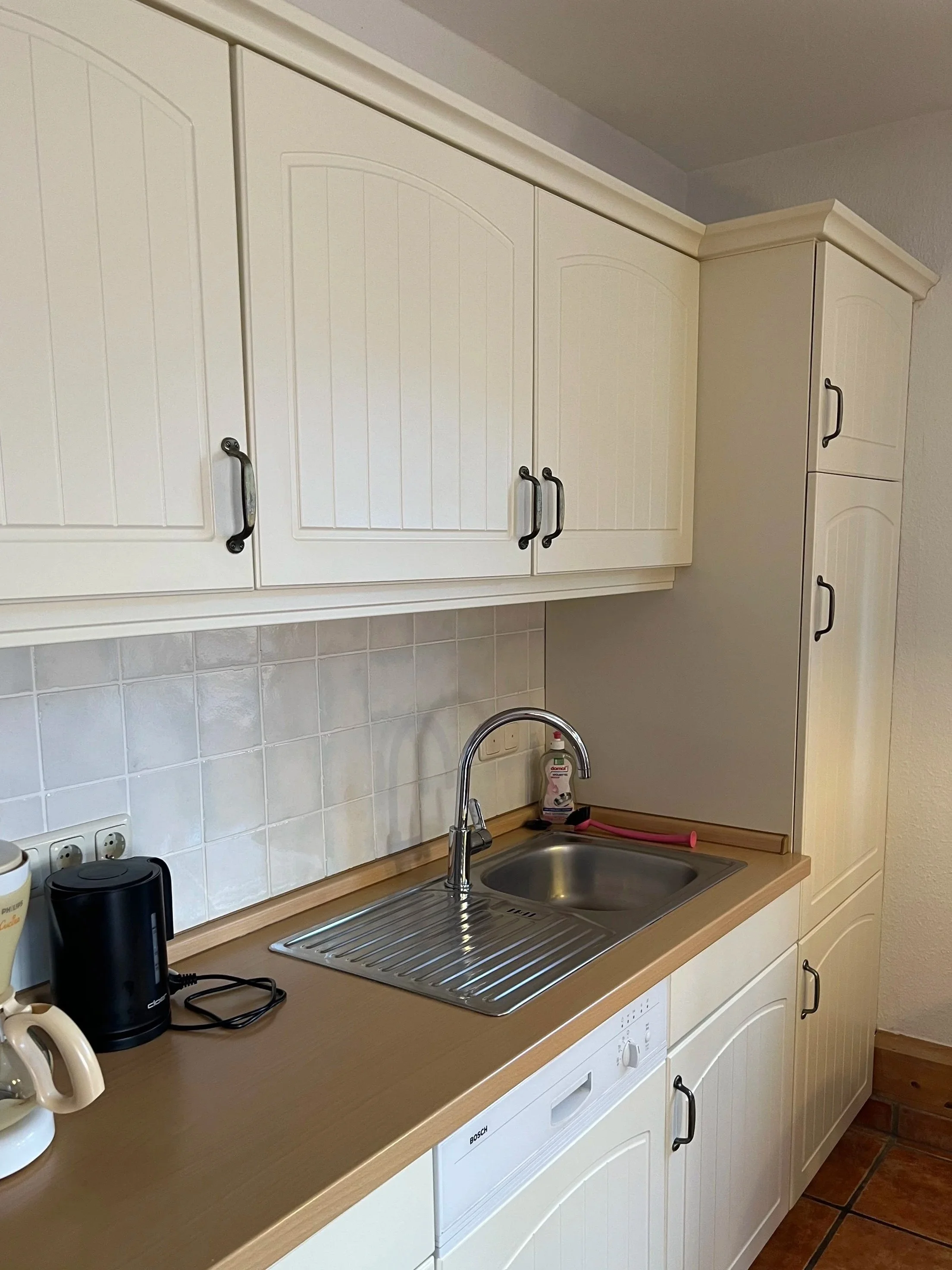 Küche mit weißen Schränken, Edelstahlspüle, beige Arbeitsplatte, schwarzen Griffen, Wasserhahn, Kaffeemaschine, Wasserkocher, Waschmaschine, white Flies backslash Küche.