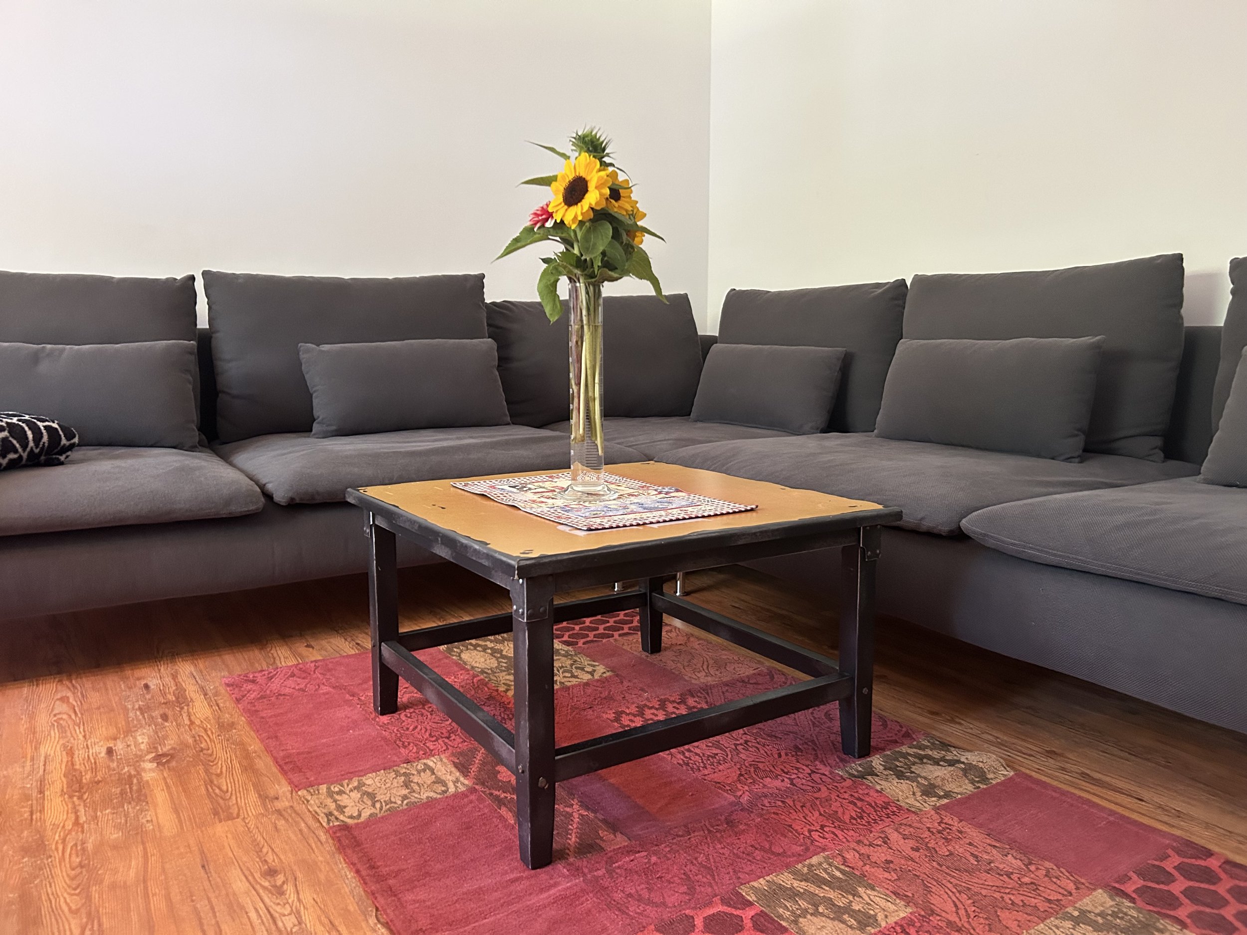 Wohnzimmer mit grauen Sofas, einem Holztisch, einem roten Teppich, einem Blumenstrauß mit Sonnenblumen auf dem Tisch, weiße Wände.