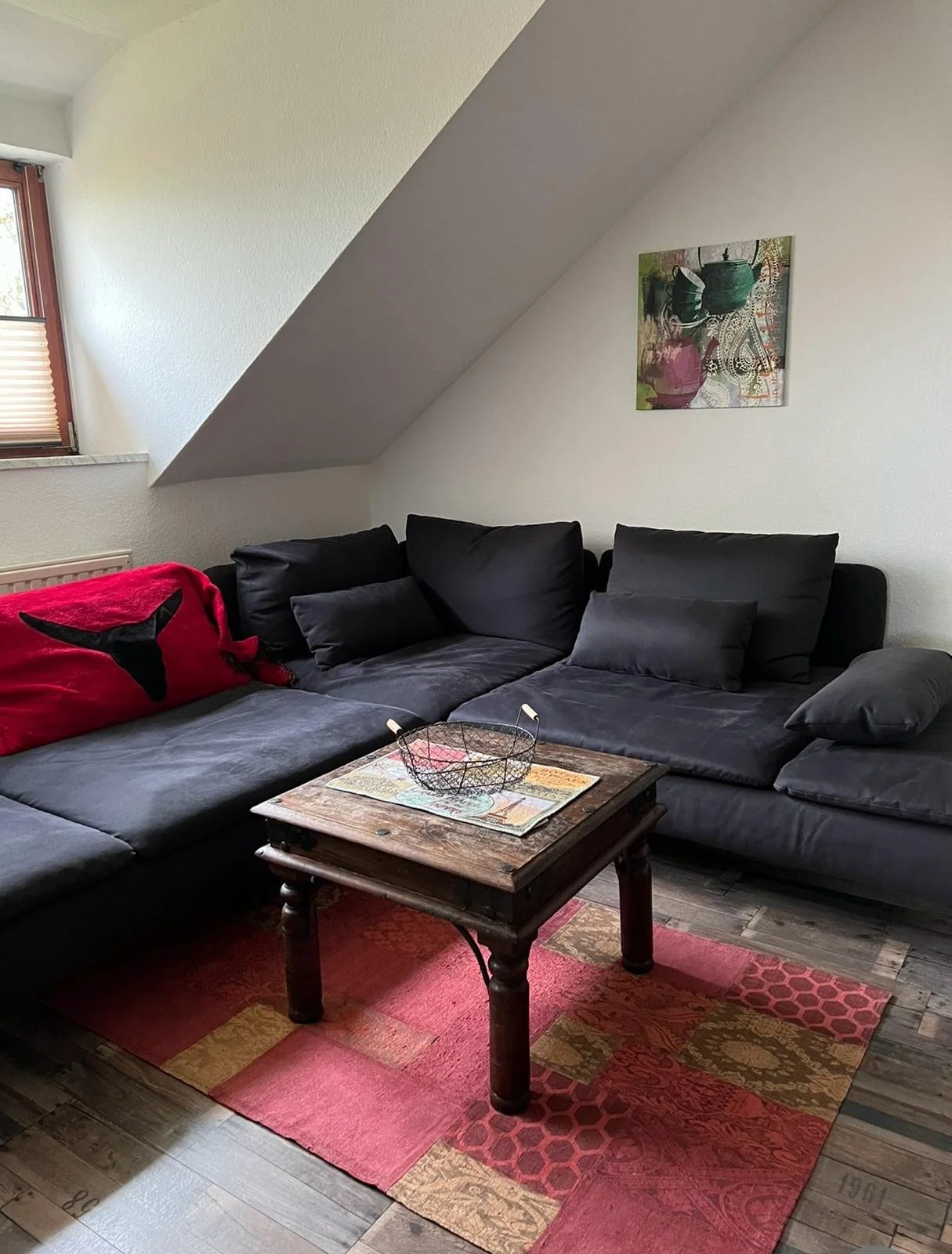 Ein Wohnzimmer mit schwarzem Sofa, einem kleinen Holztisch, einem bunten Teppich, einem Wandbild und einem Fenster mit Rolladen unter einer Dachschräge.