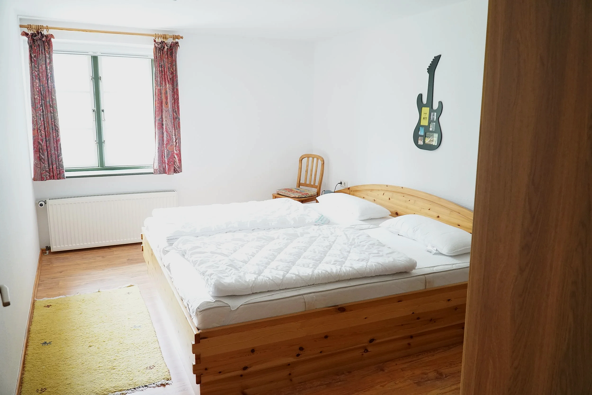 Zimmer mit Doppelbett, Fenster mit roten Vorhängen, Holzfußboden, Wanddeko in Form einer Gitarrenform, Stuhl mit Kissen, Matratzen und Bettwäsche.