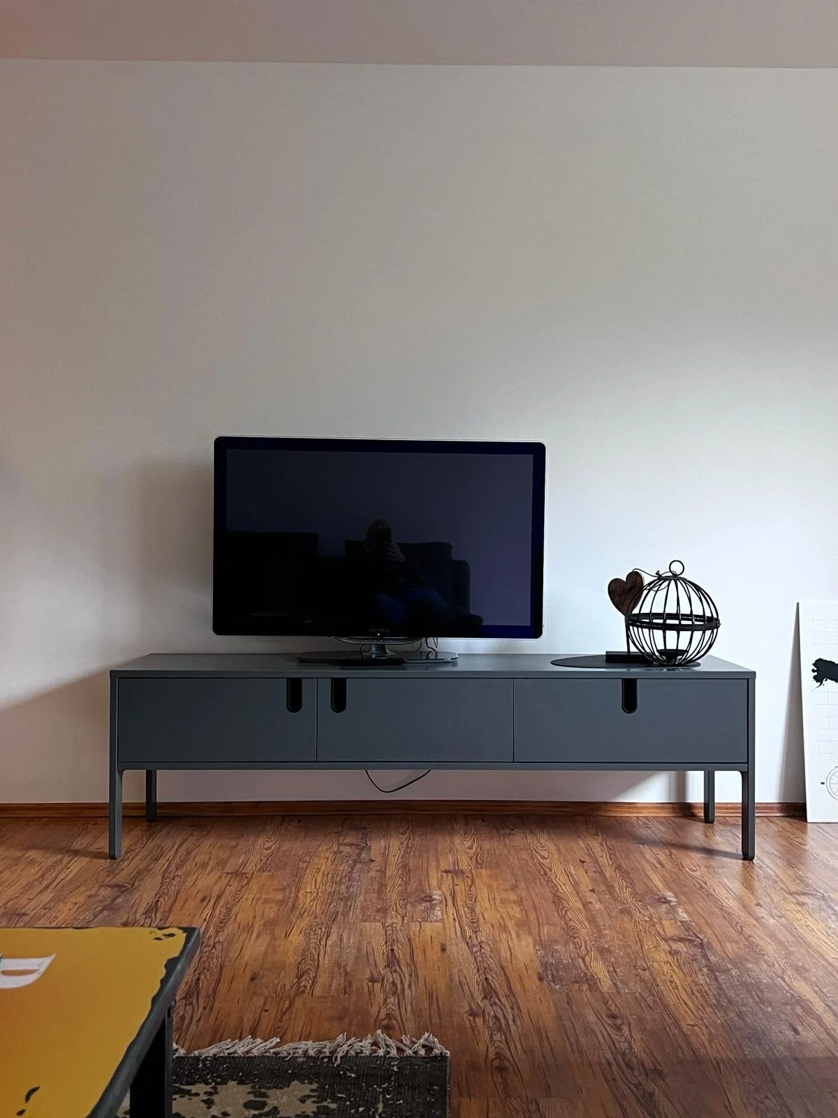 Moderner Fernsehtisch mit schwarzem Fernseher und dekorativen Gegenständen auf einem Holzboden, weiße Wand im Hintergrund.