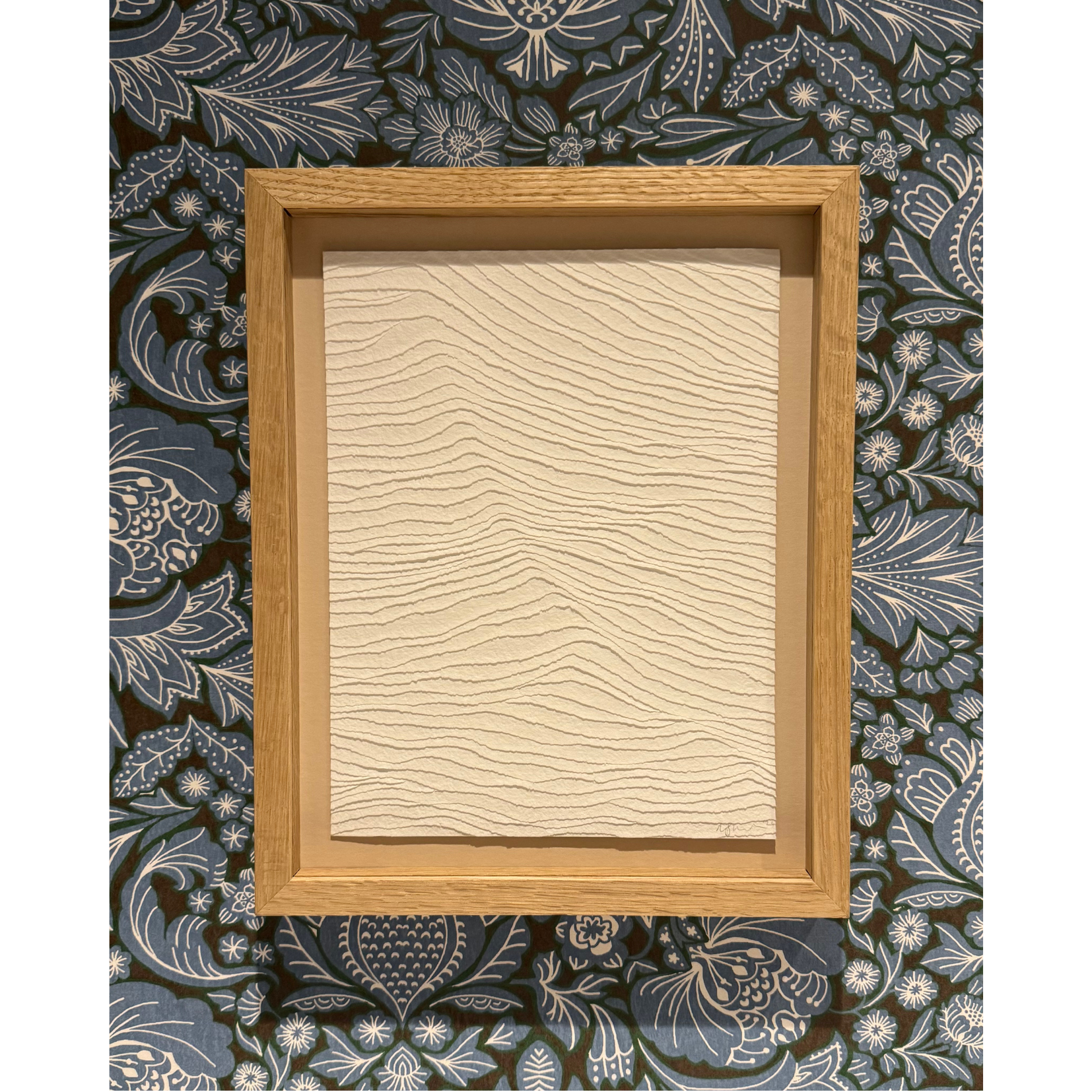 white textural artwork of gentle waves in the sea. Framed in oak and shown against a patterned blue wallpaper