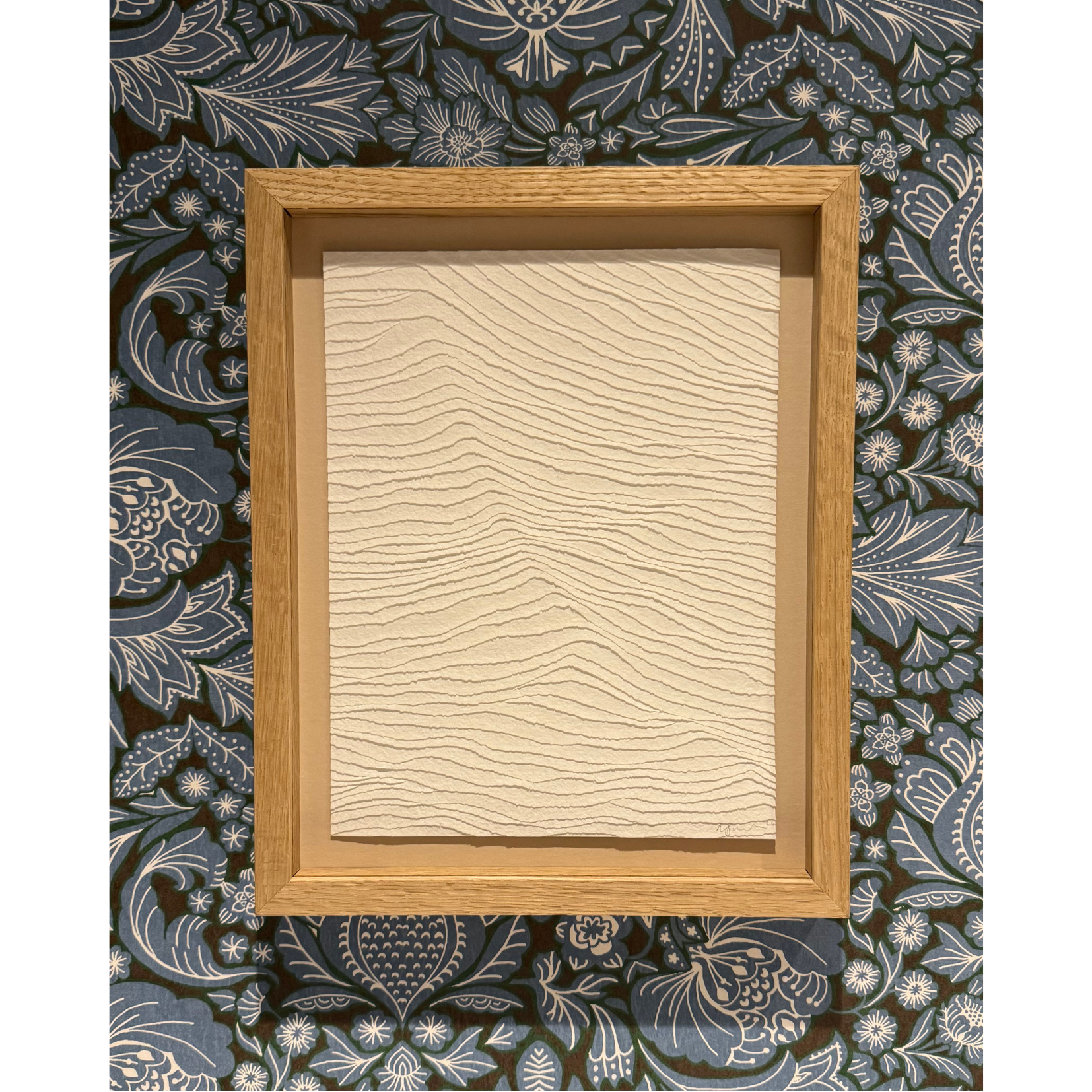 white textural artwork of gentle waves in the sea. Framed in oak and shown against a patterned blue wallpaper