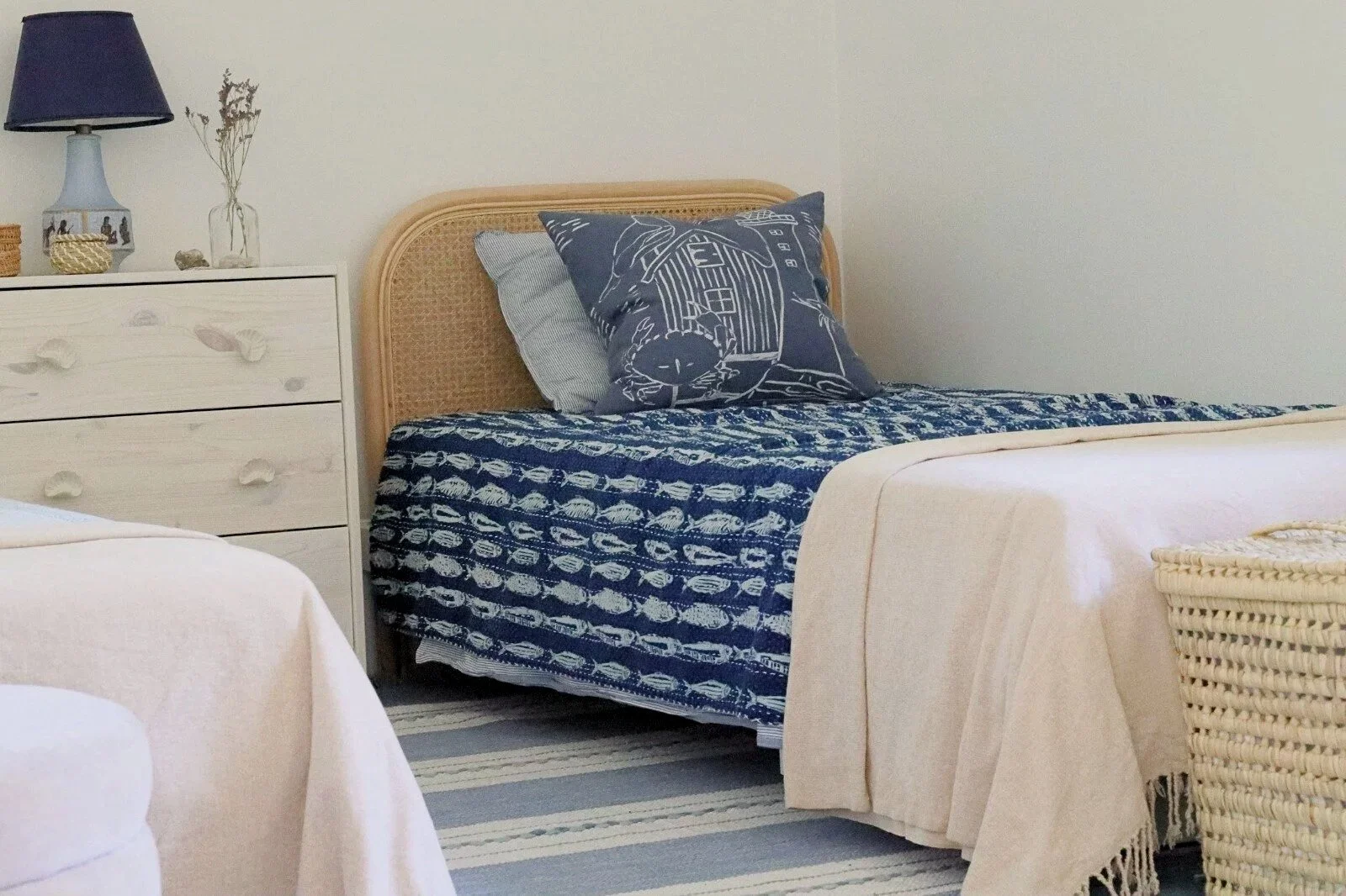 Enkelsäng med rundad rottinggavel klädd med ett marinblått fiskmönstrat överkast och en persikofärgad filt, med en marinblå kustmönstrad kudde. Bredvid sängen står en vitlackad furubyrå med musselsformade draglådknopp, dekorerad med en marinblå bords