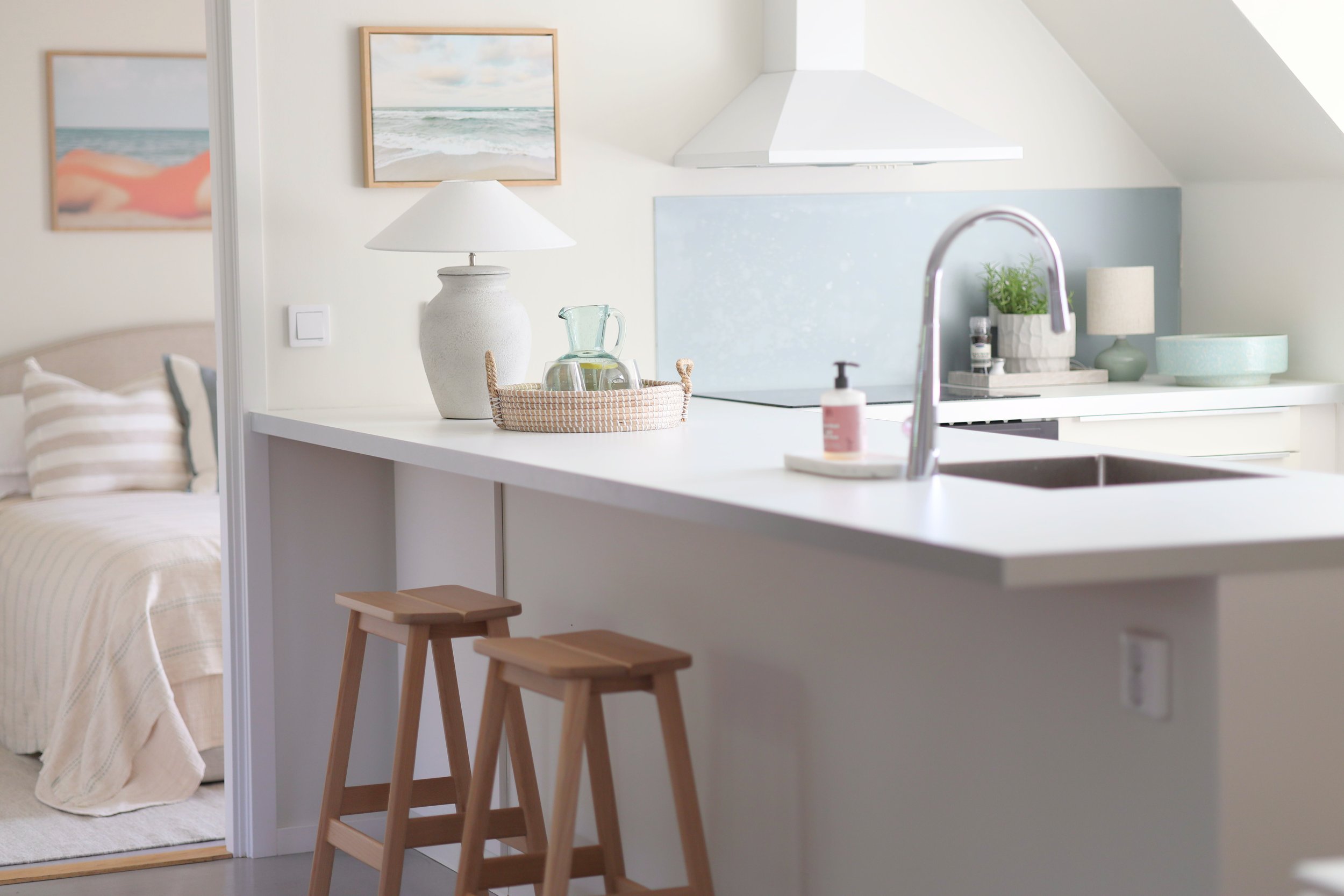 Kök med vita skåp, diskho och kran, några gröna växter och ljus inredning. Strykningslampor och tavlor på väggarna.