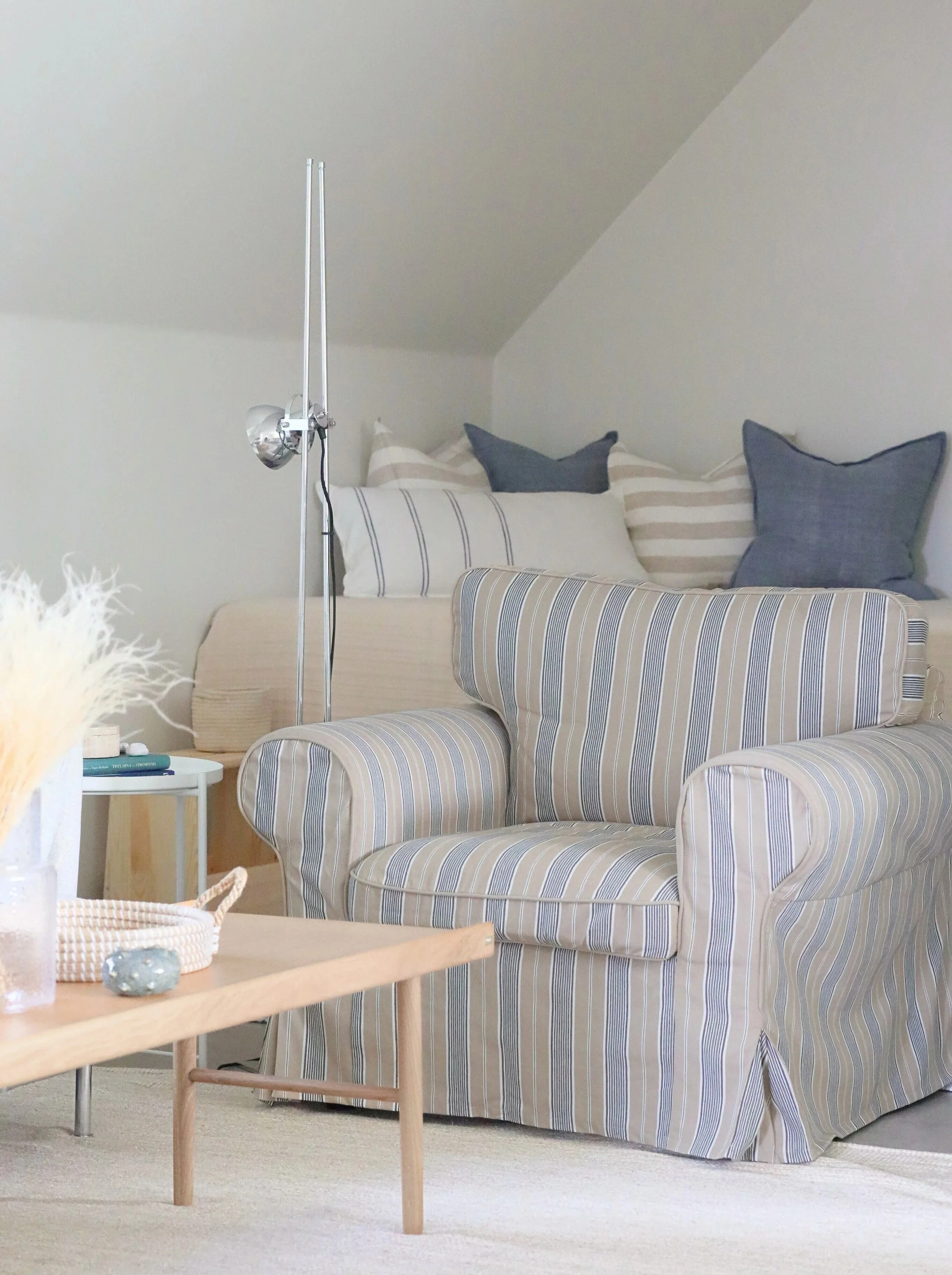 En liten soffa med randigt tyg i beige och blått framför en vägg med en grå soffa eller säng med blå och vita kuddar. I förgrunden syns ett lågt bord med dekorationer.