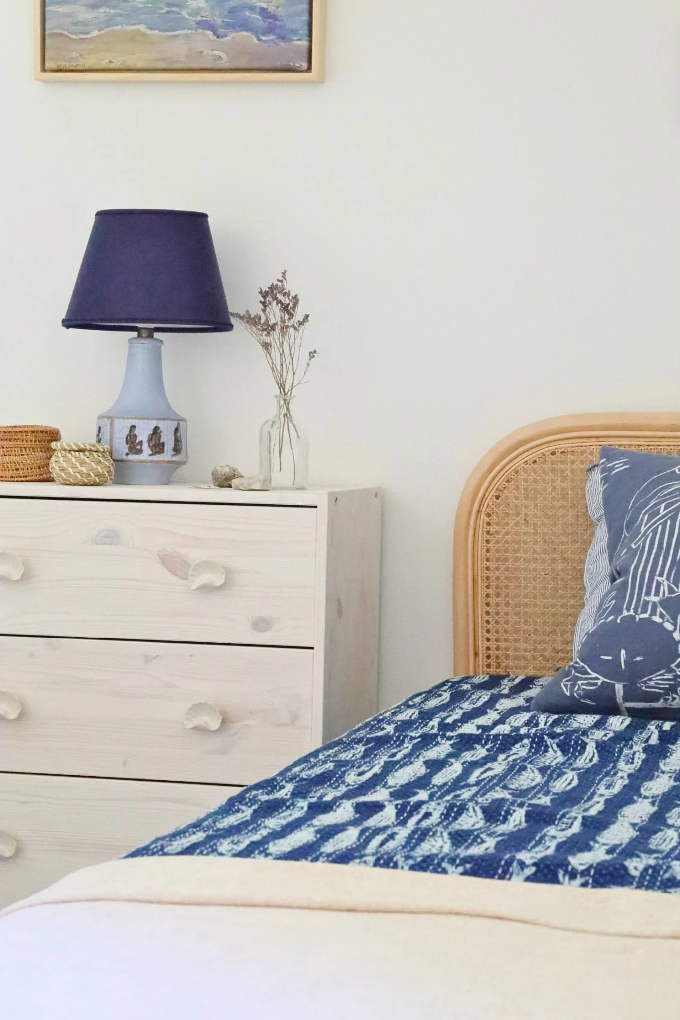 Enkelsäng med rundad rottinggavel klädd med ett marinblått fiskmönstrat överkast och en persikofärgad filt, med en marinblå kustmönstrad kudde. Bredvid sängen står en vitlackad furubyrå med musselsformade draglådknopp, dekorerad med en marinblå bords
