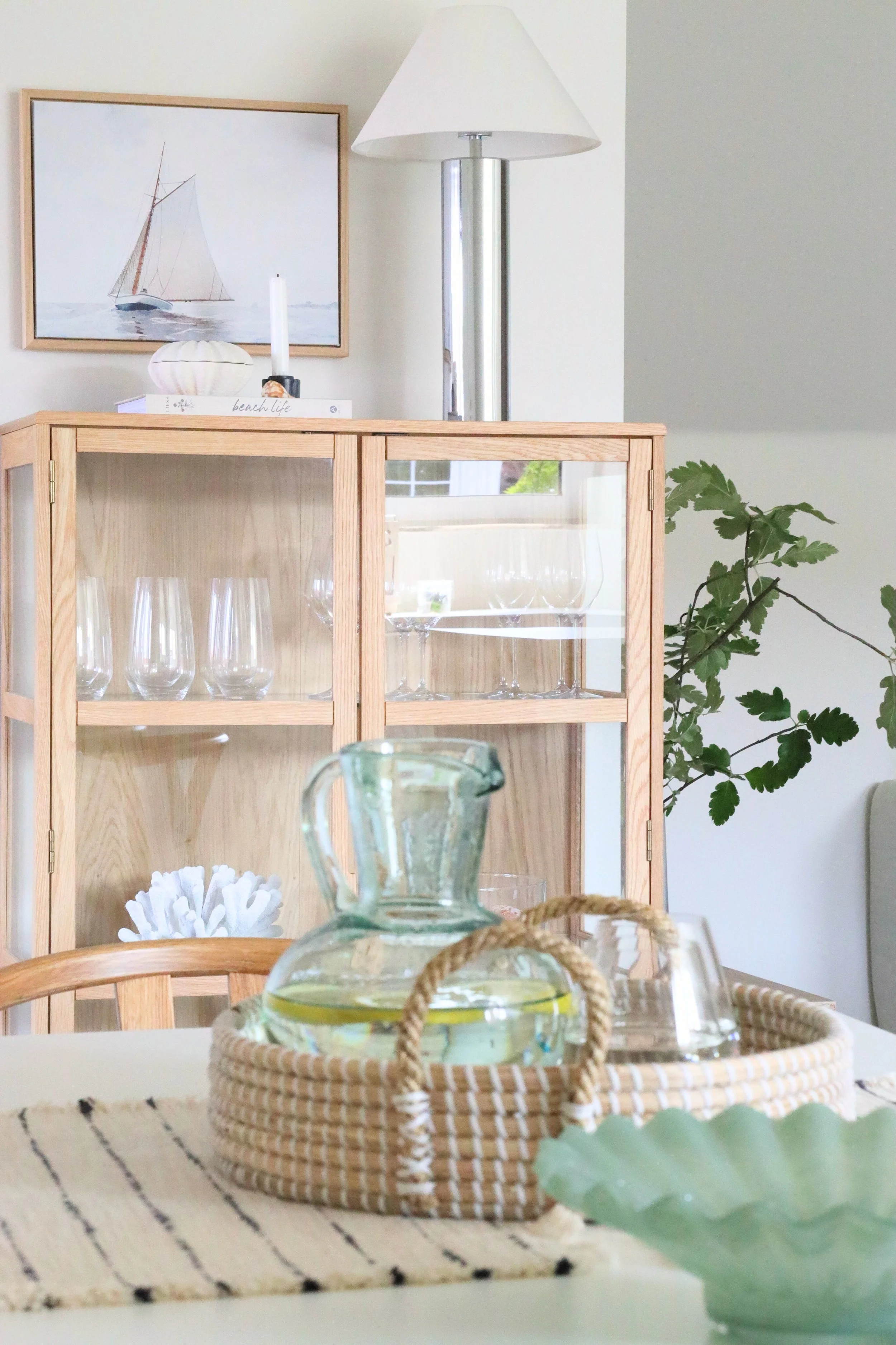 Ett vardagsrum med en glasvas med vatten och citron, ett fat med en kopp, en glasvas, en soffa och gröna växter. I bakgrunden ett skåp med glas och en väggdekoration med motiv av en segelbåt.