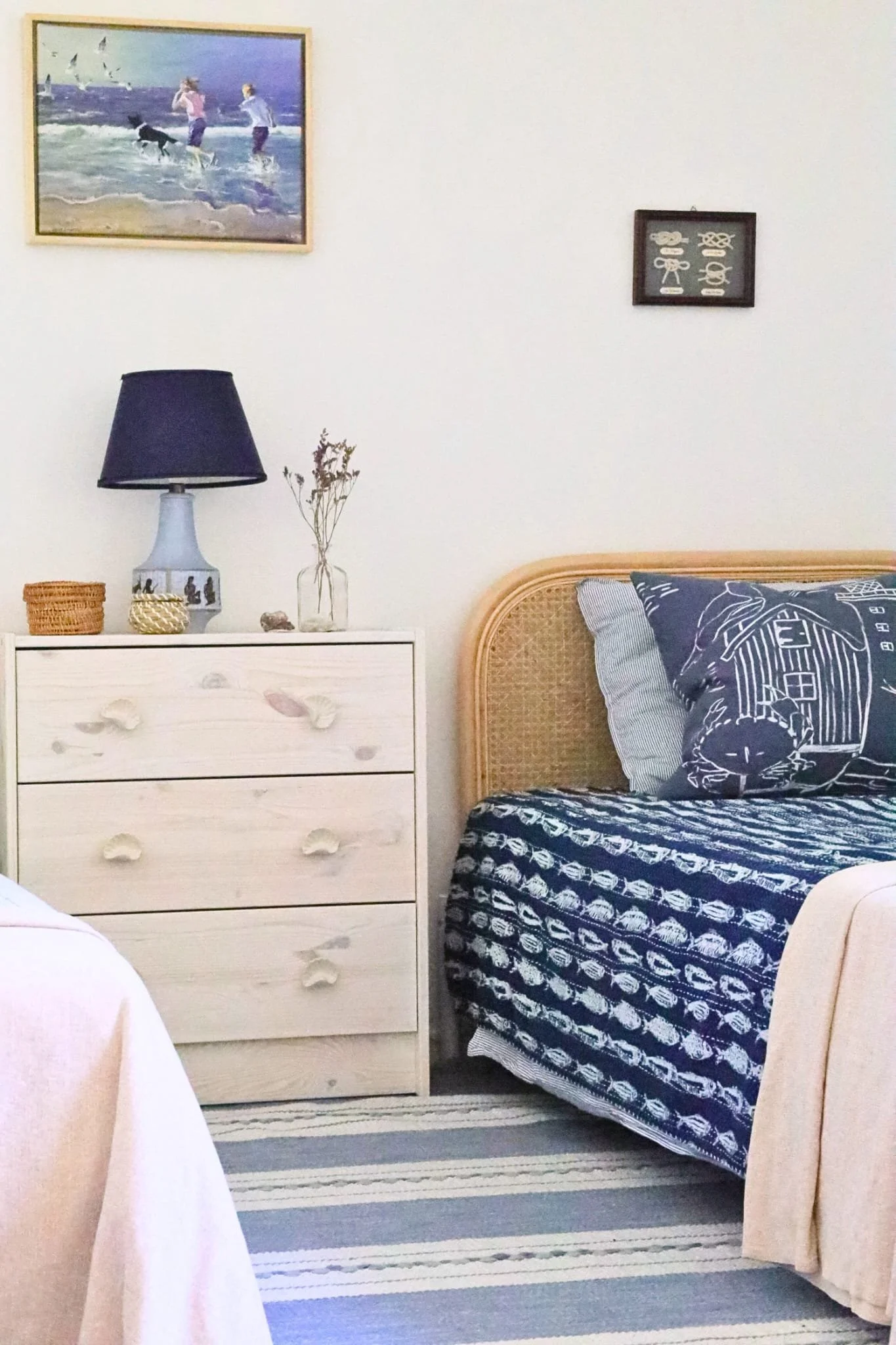 Delat barnrum fotograferat från motsatt vinkel. En enkelsäng med rundad rottinggavel klädd med ett marinblått fiskmönstrat överkast och en persikofärgad filt, med marinblå kustmönstrade kuddar. Mellan sängarna står en vitlackad furubyrå med musselsfo