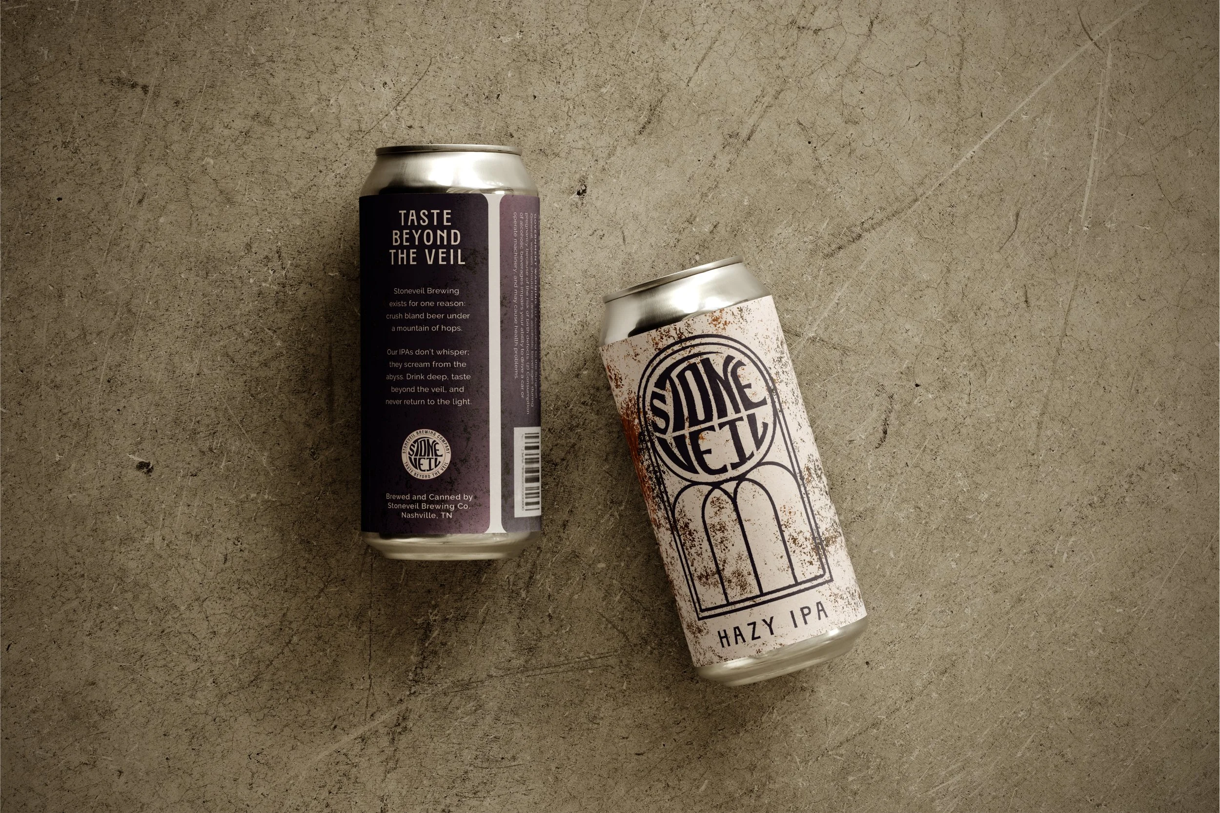 Two cans of craft beer on a concrete surface, one labeled "Taste Beyond The Veil" and the other labeled "Hazy IPA" with a black and white geometric design.