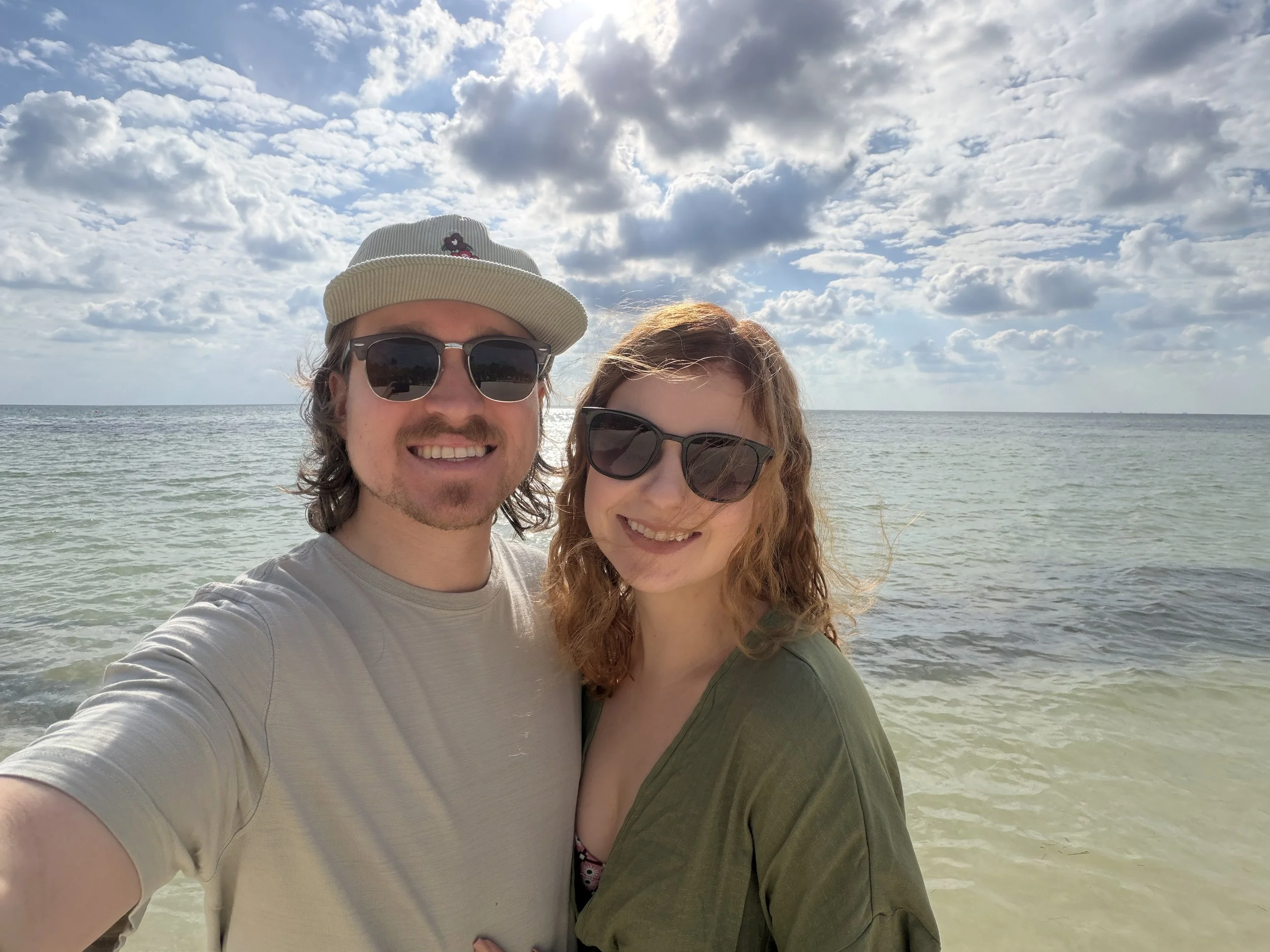 A smiling man and woman wearing sunglasses taking a selfie at the beach with ocean and cloudy sky in the background.