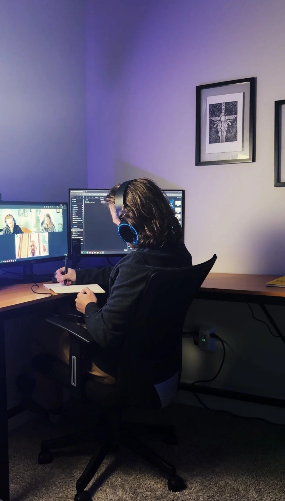 A person with long, dark hair is sitting at a desk with two computer monitors, wearing large headphones, and taking notes. The monitors display video conferencing screens and a desktop with various folders and icons. The room has purple lighting, framed artwork containing cross images on the wall, and a power strip on the floor.