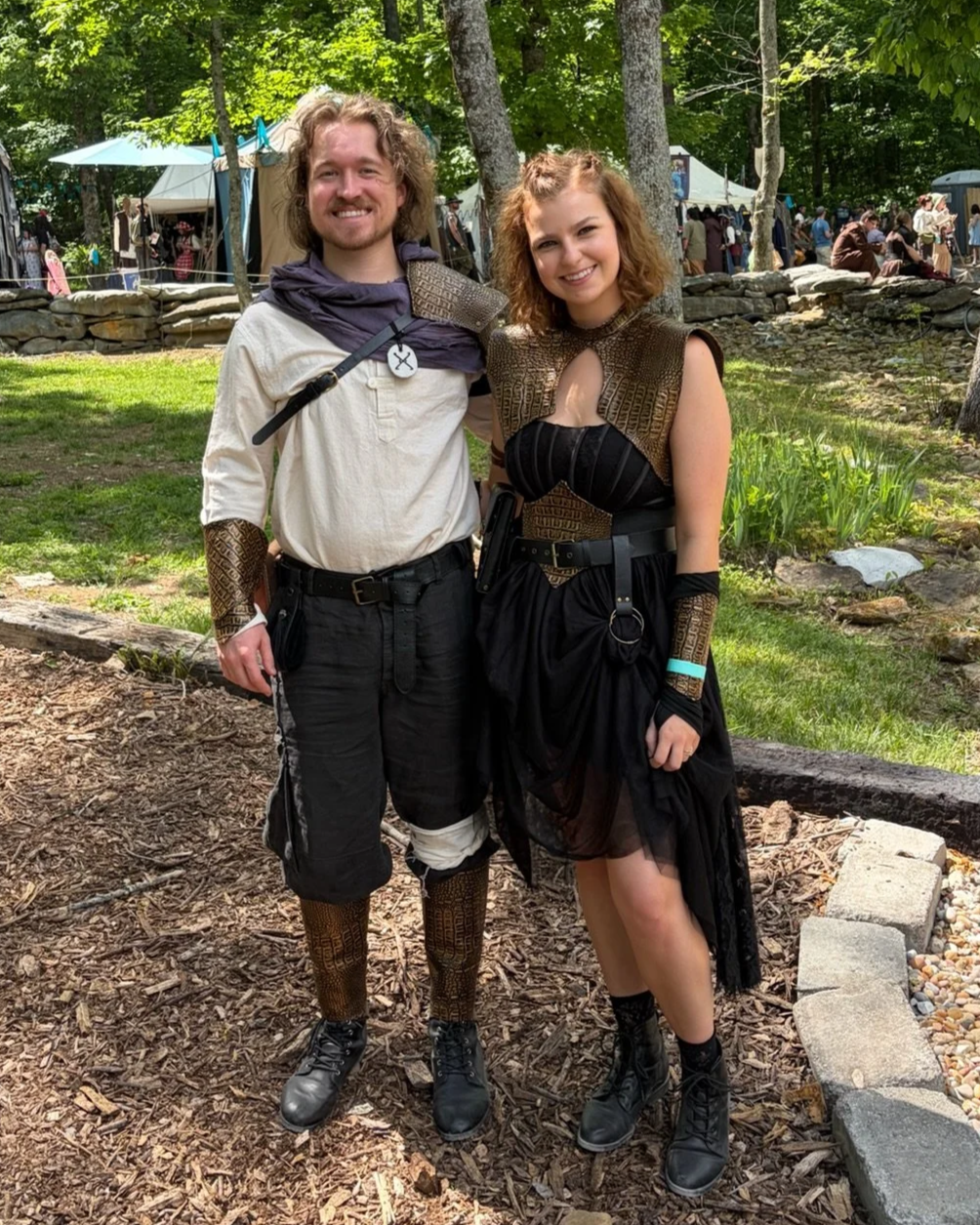 A man and woman dressed in medieval or fantasy costumes, standing outdoors on a dirt path with trees and festival tents in the background.