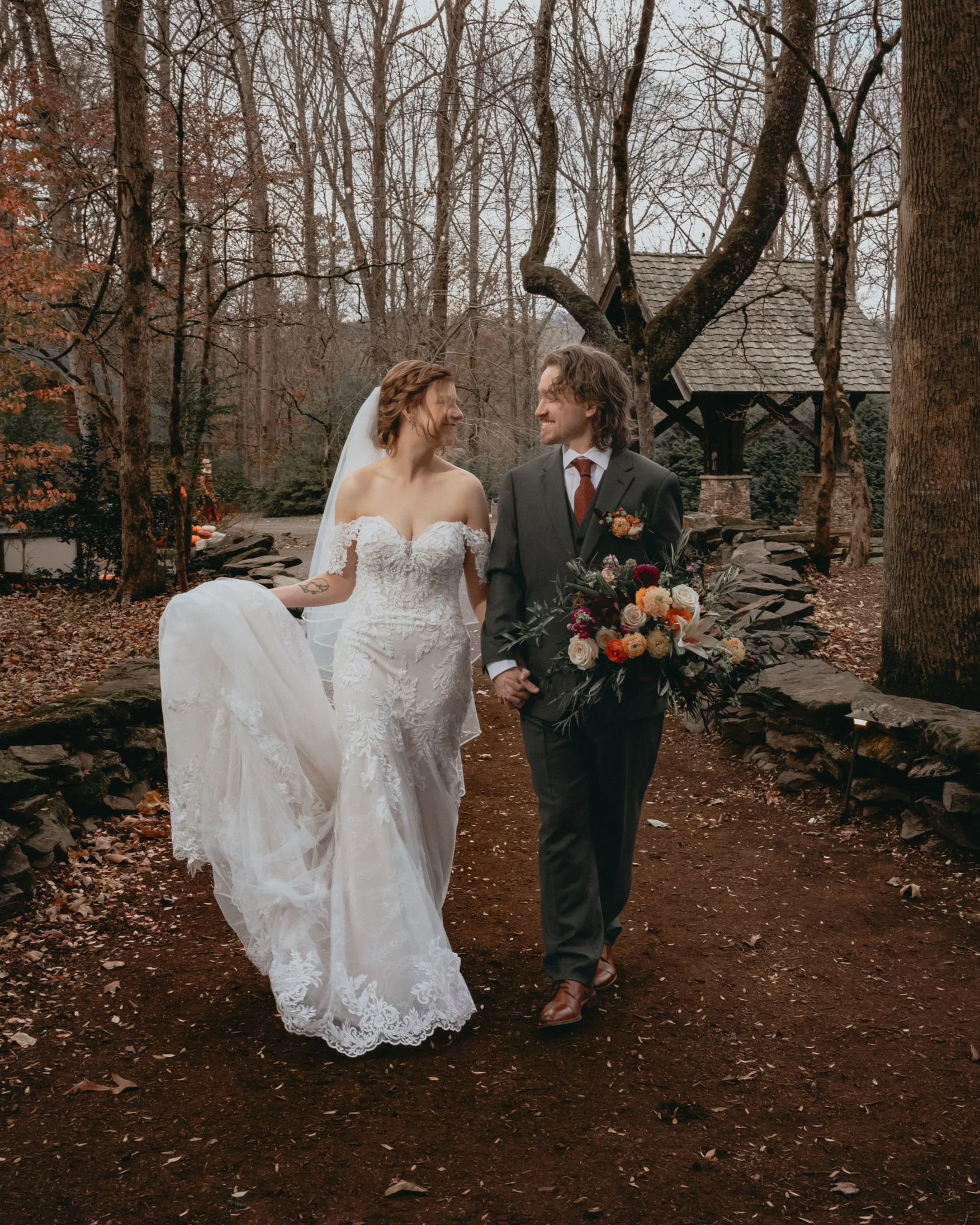 A bride and groom happily holding hands and smiling at each other during their wedding in an outdoor wooded area with trees, fallen leaves, and a small stone well in the background.