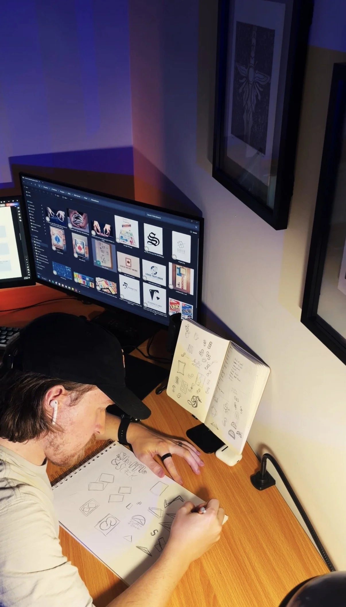 Young man drawing geometric shapes and logo concepts on paper in an office, with a computer monitor showing design thumbnails, documentation, and framed artwork on the wall.