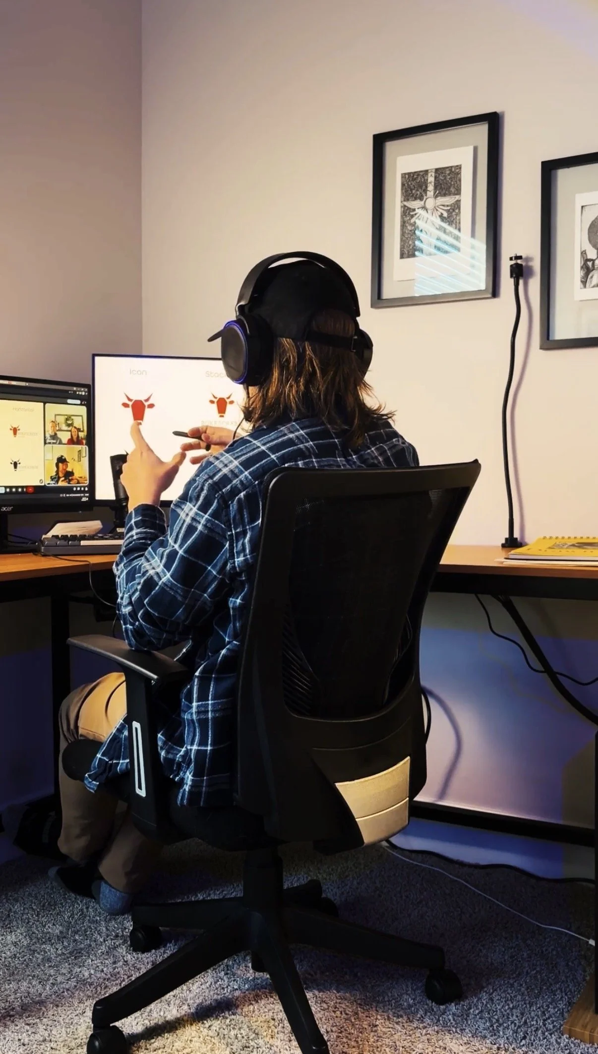 Person wearing headphones sitting at a desk with two computer monitors, one displaying a logo of a red dragon head, and the other with video call windows. The person is in a room with artwork on the wall.