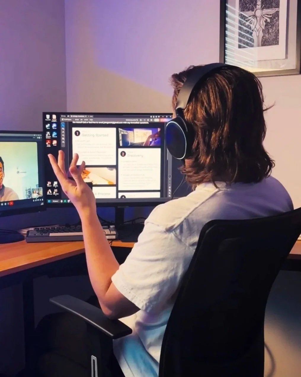 Person sitting at a desk with two computer monitors, wearing large over-ear headphones, and gesturing with their hand while participating in a video call or virtual meeting.