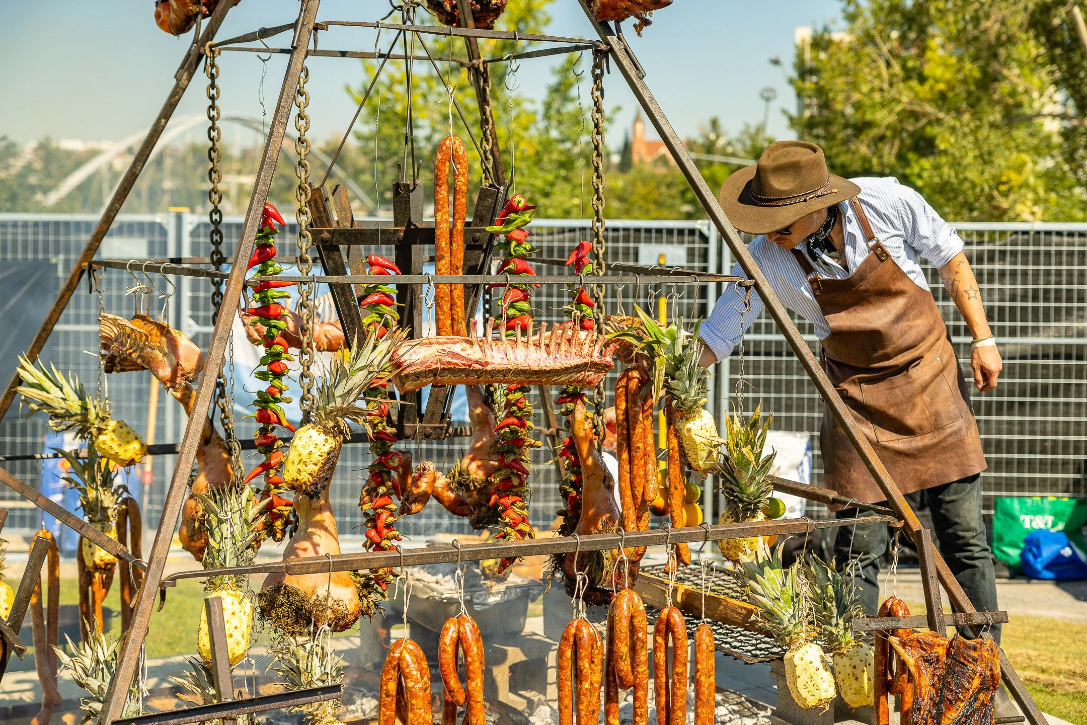 A person wearing a large hat, sunglasses, a striped shirt, and a brown apron is tending to a barbecue grill with various meats, pineapples, and vegetables hanging from a metal frame. The scene takes place outdoors on a sunny day.