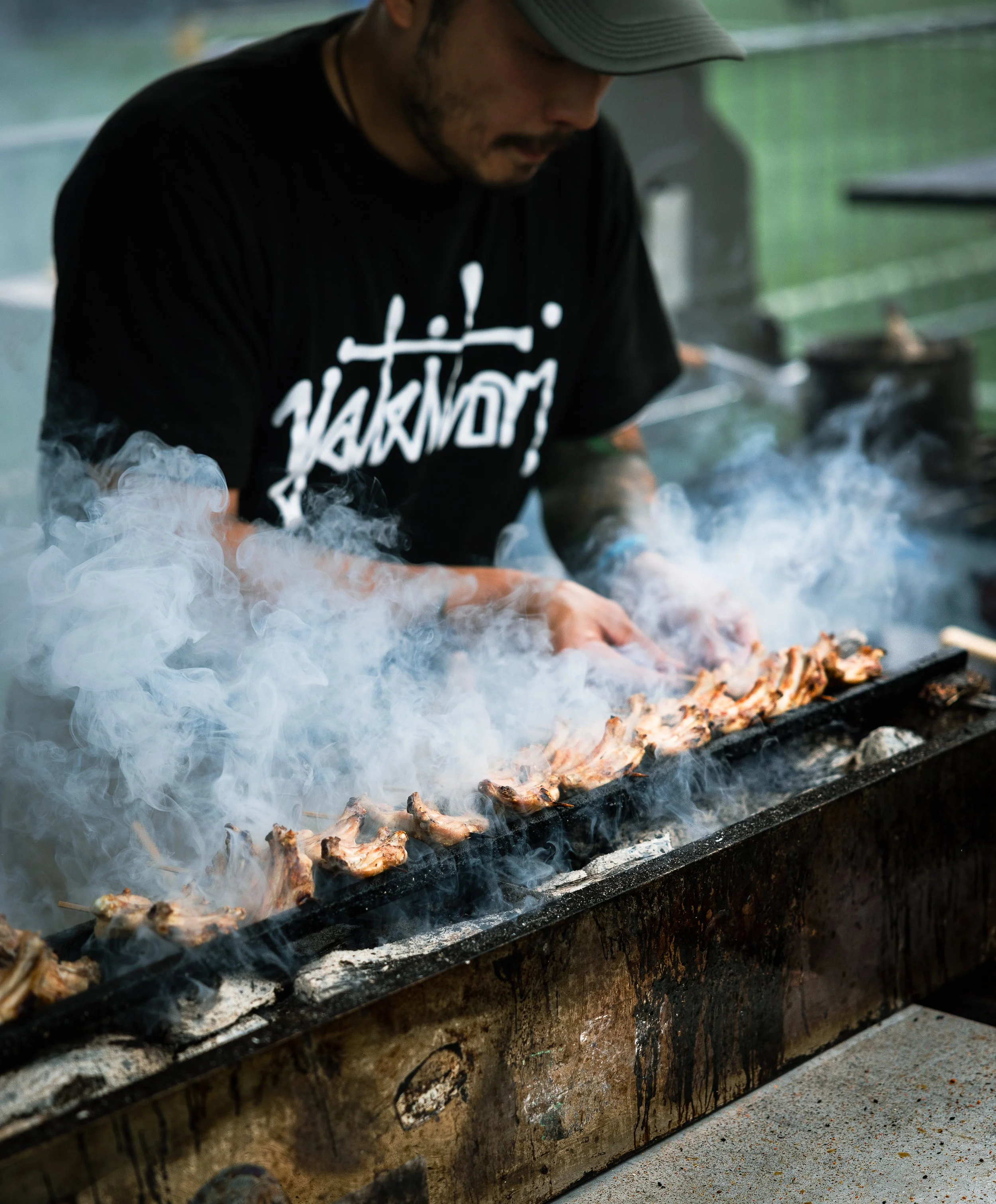 A man wearing a black t-shirt and hat grilling skewers of meat on a smokey barbecue grill.