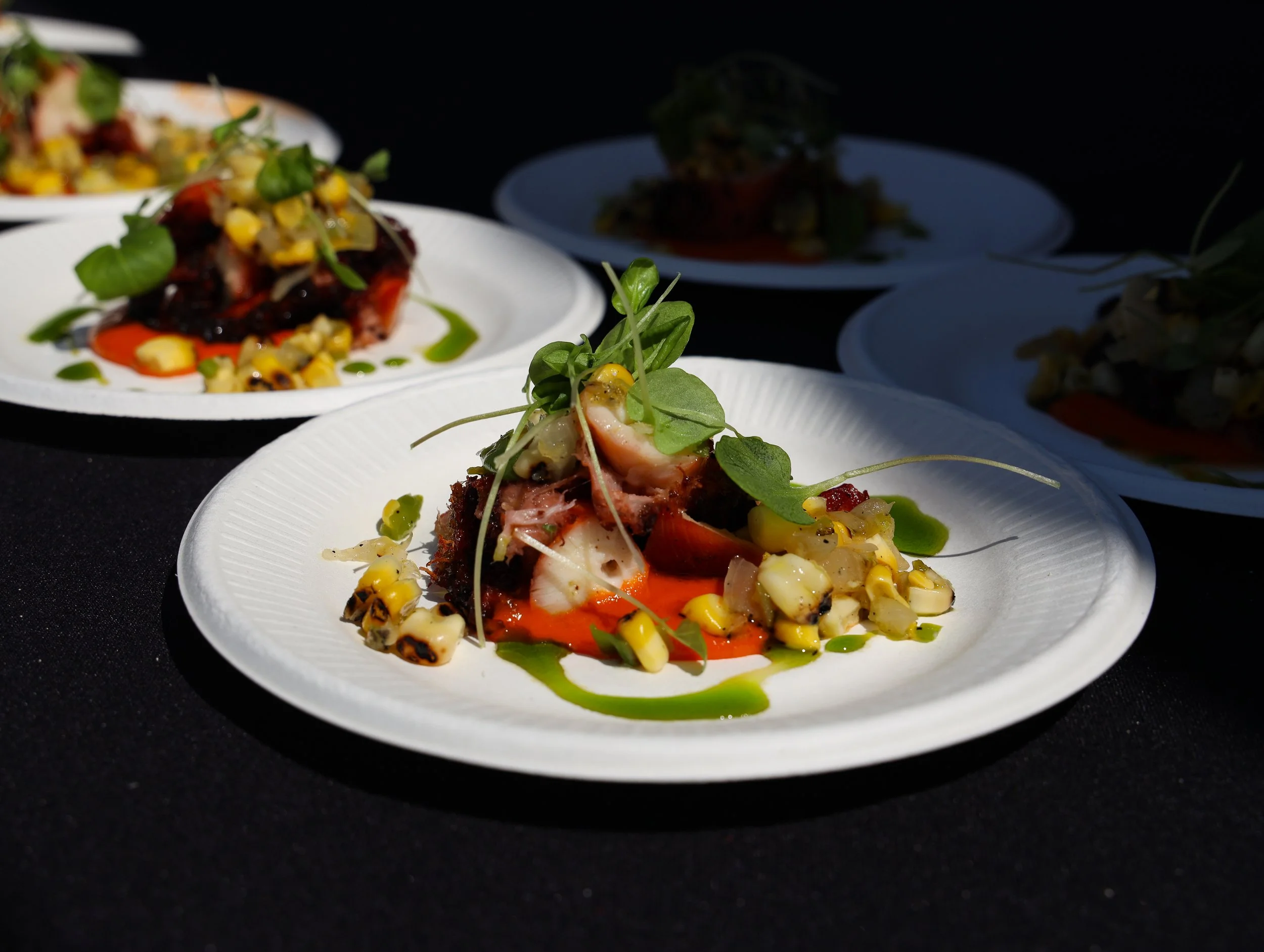 A close-up of a plated gourmet dish featuring grilled or roasted meat with microgreens, corn, and a red sauce on a white plate against a dark background.