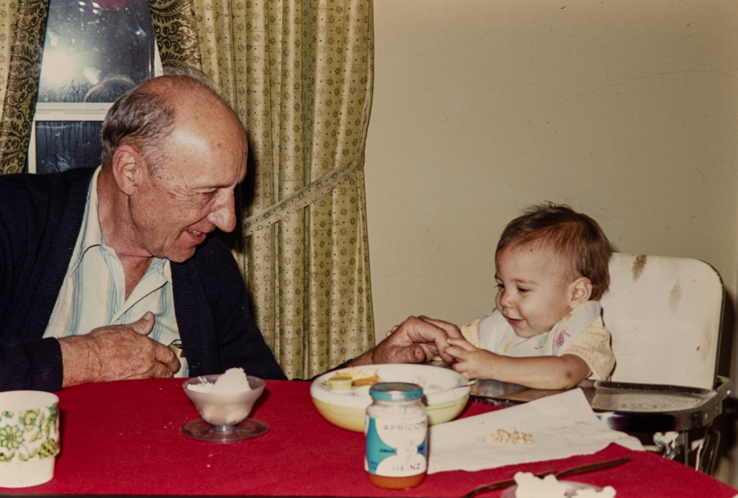 An elderly man and a young girl are sitting at a table, holding hands and smiling at each other. The table has bowls of food and medicine bottles. They are in a room with patterned curtains and a window.