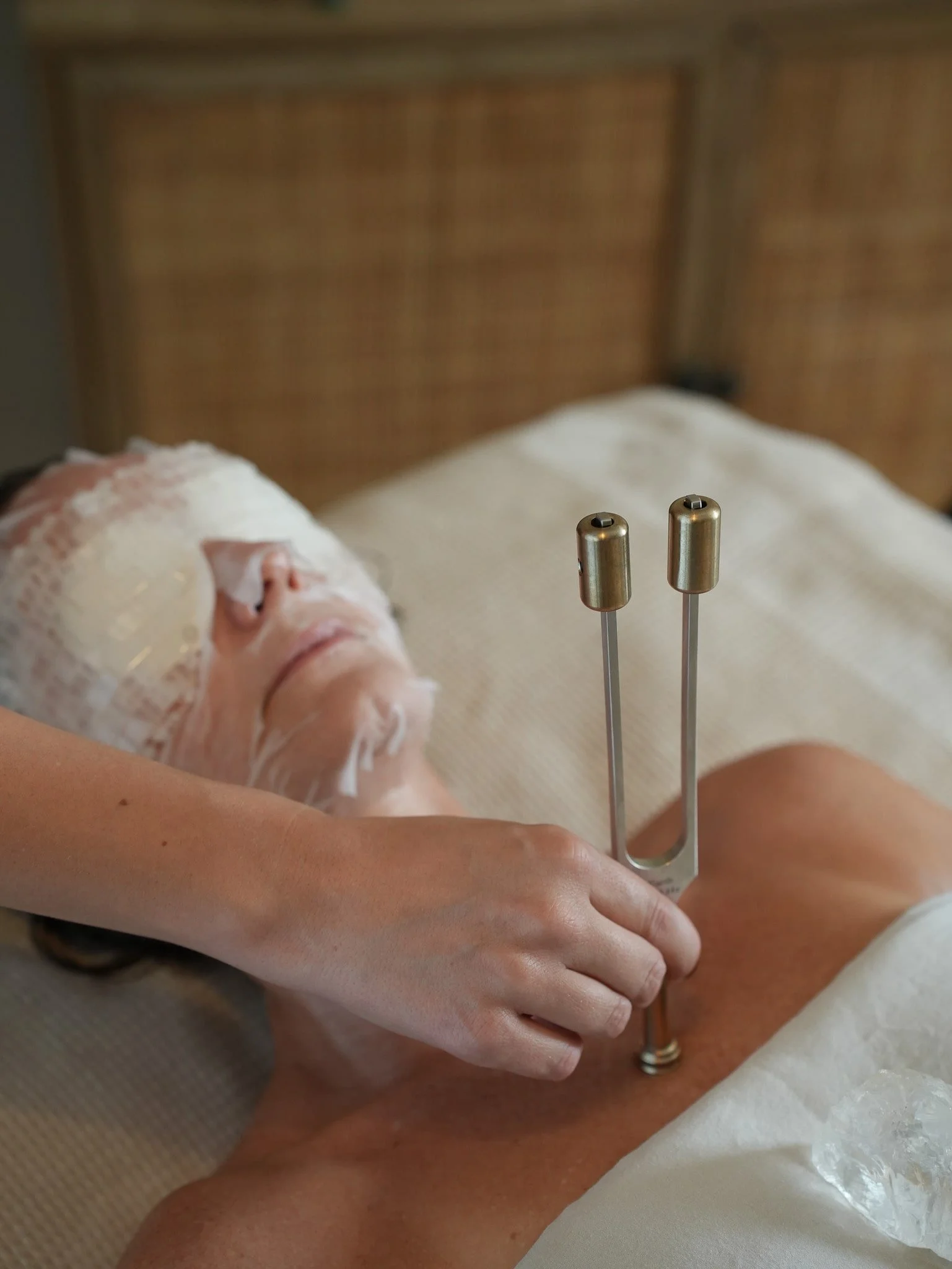 A woman lying in a spa bed receiving a treatment with two metal acupuncture-like needles inserted into her stomach, with her face covered in a white facial mask.