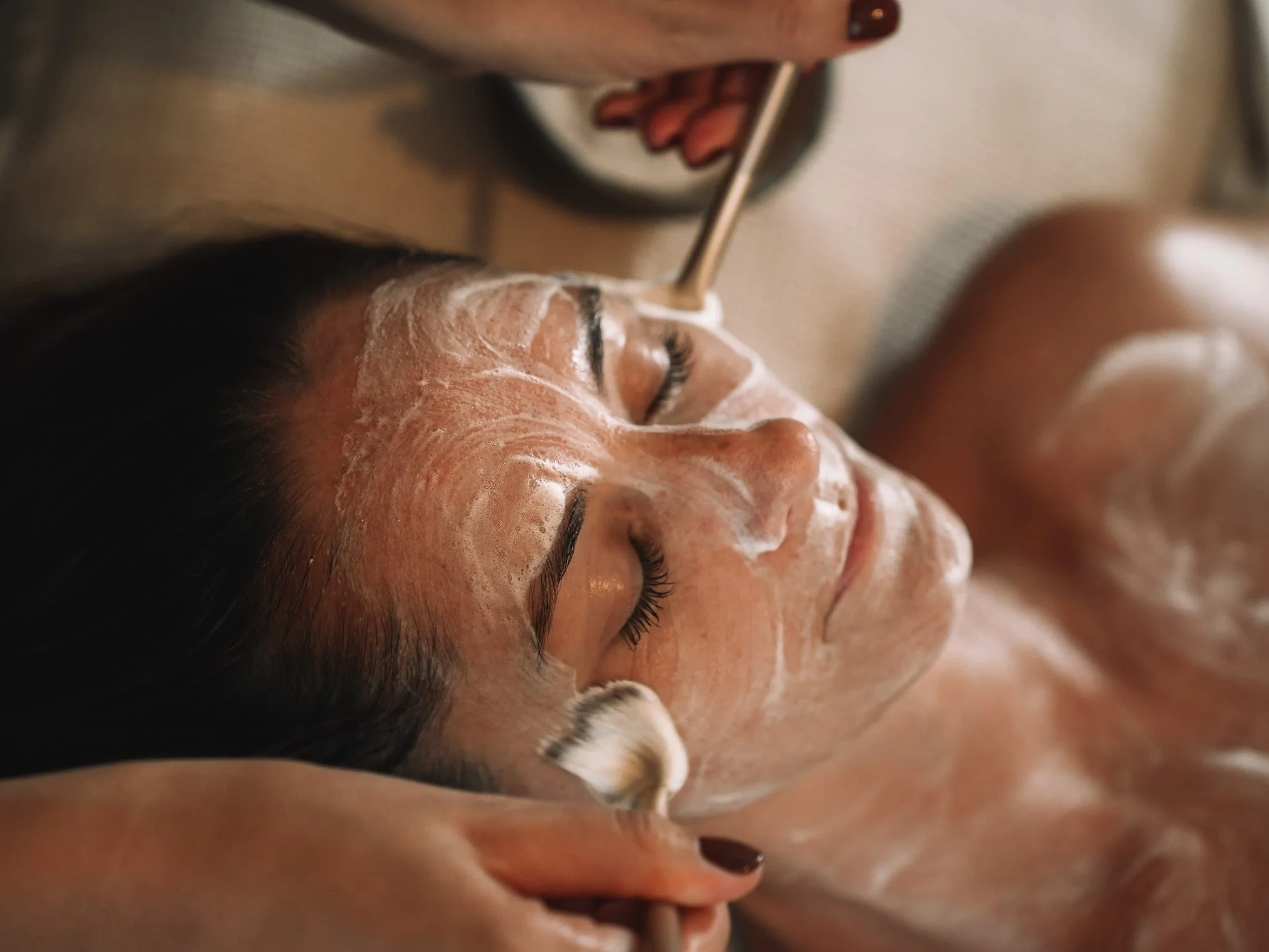 A woman receiving a facial treatment, lying on a spa bed with her eyes closed. A skincare product is applied to her face.