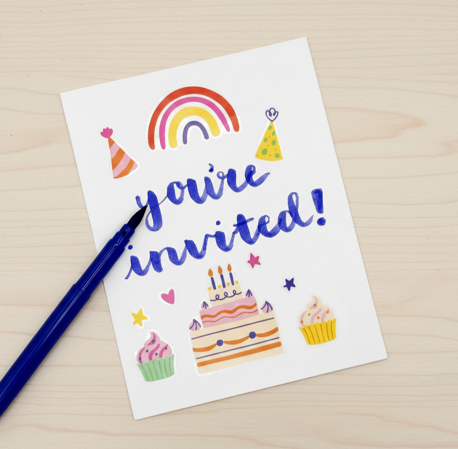 A colorful birthday card on a light wood surface with the words "you're invited!" written in blue, decorated with a rainbow, party hats, cupcakes, a birthday cake with candles, and small stars and hearts.