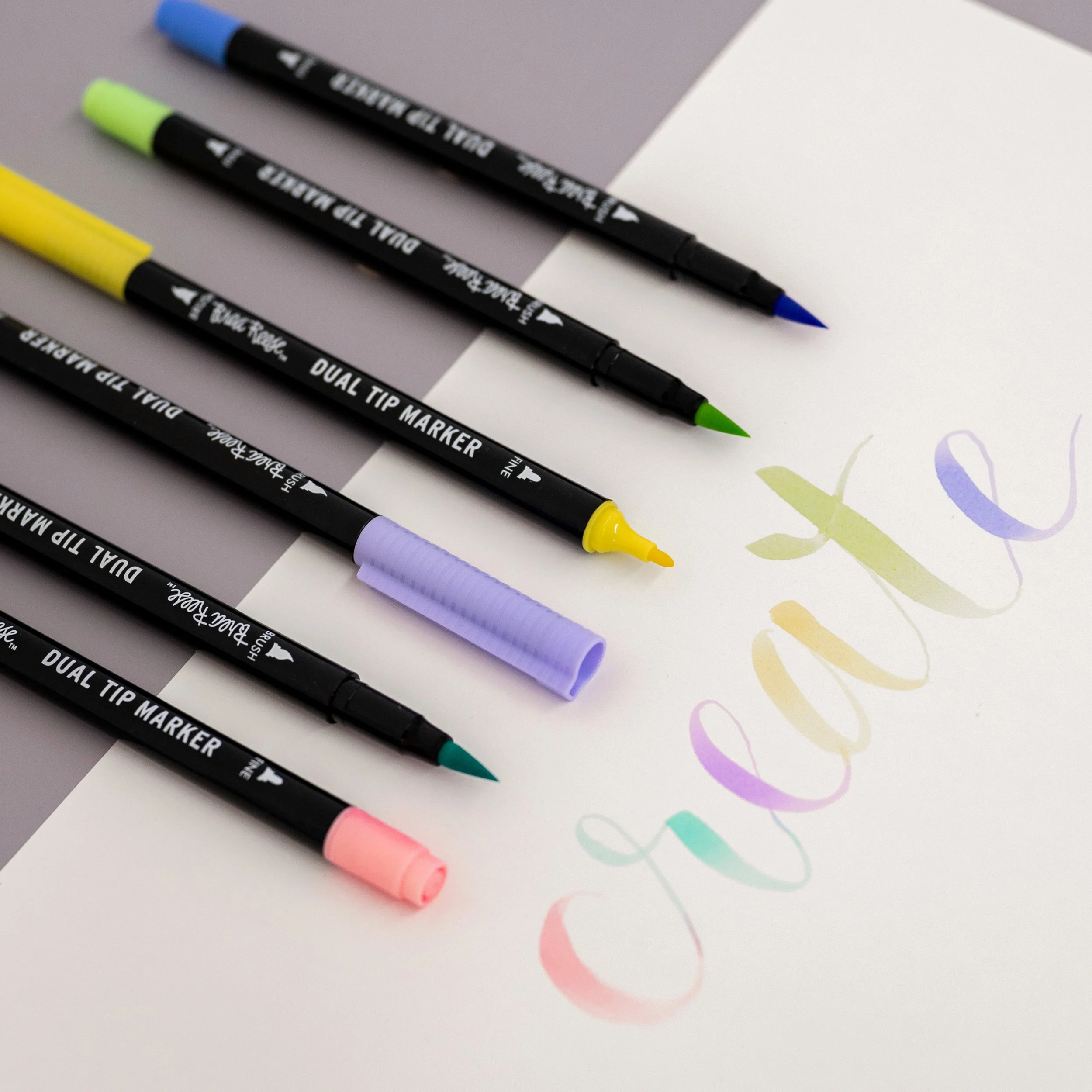 Five dual tip markers in various pastel colors lying on paper with the word 'love' written in colorful, cursive calligraphy.