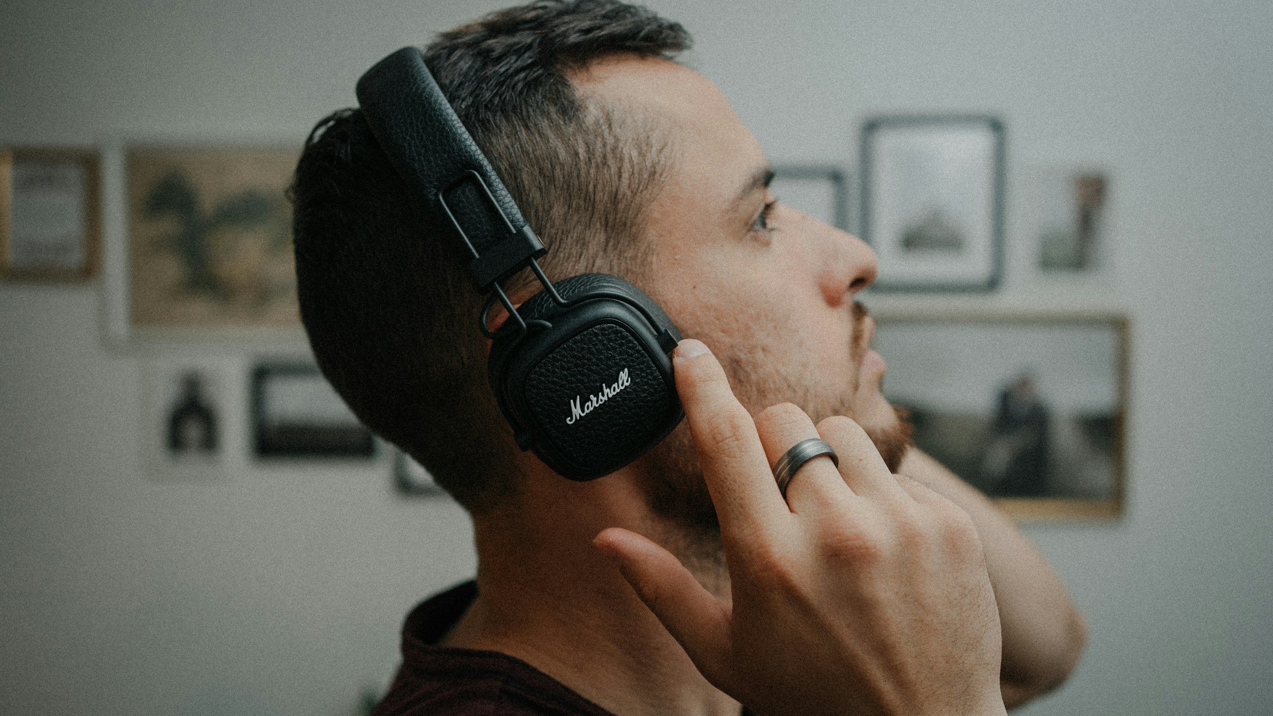 A man with short hair and a beard wearing black Marshall headphones, touching the side of the headphones with his right hand, which has a silver ring on the middle finger, in a room with framed pictures on the wall.