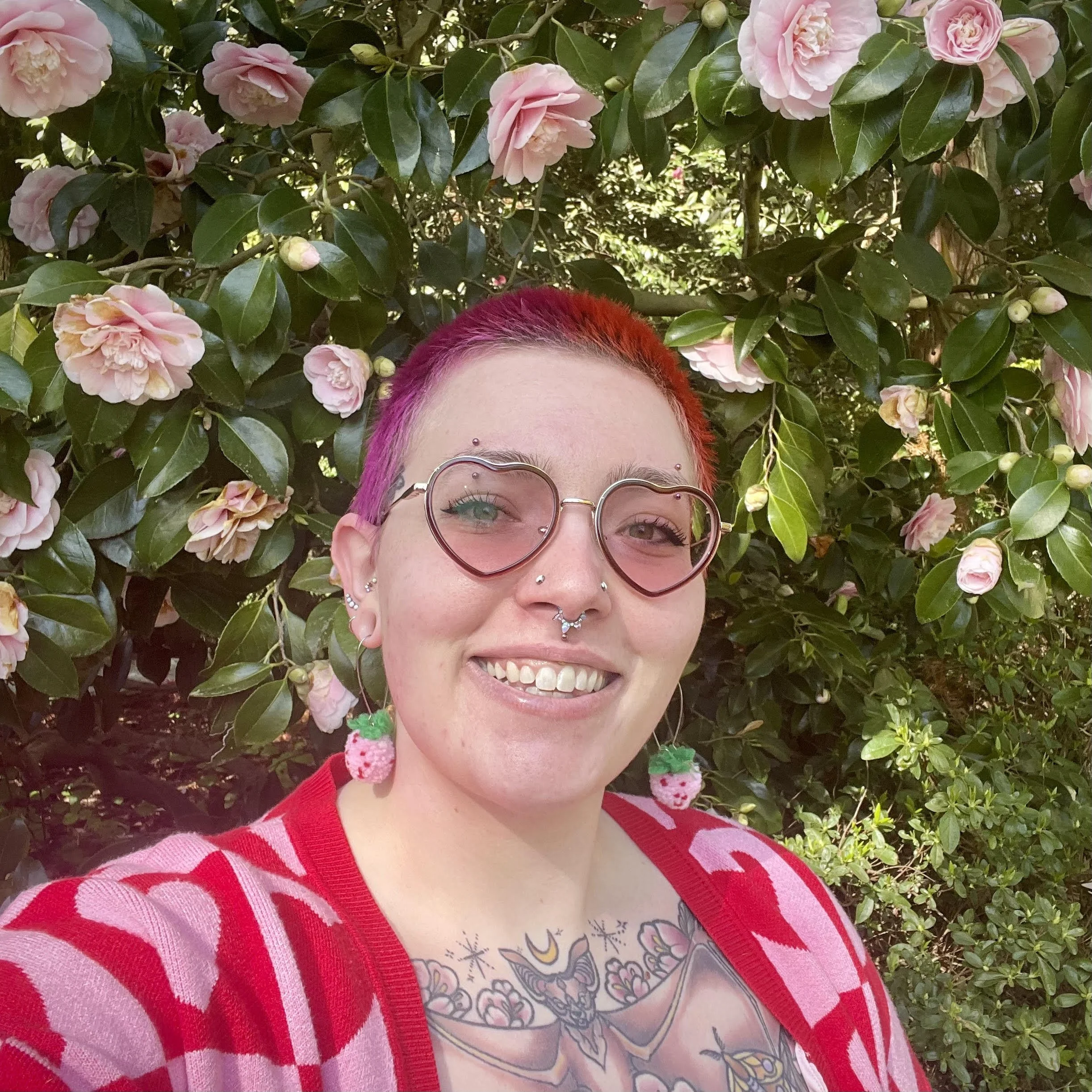 A person with short red hair smiling, wearing heart-shaped glasses, pink hoodie, and colorful earrings, with tattoos on their hand, in front of a pink wall with colorful artwork.