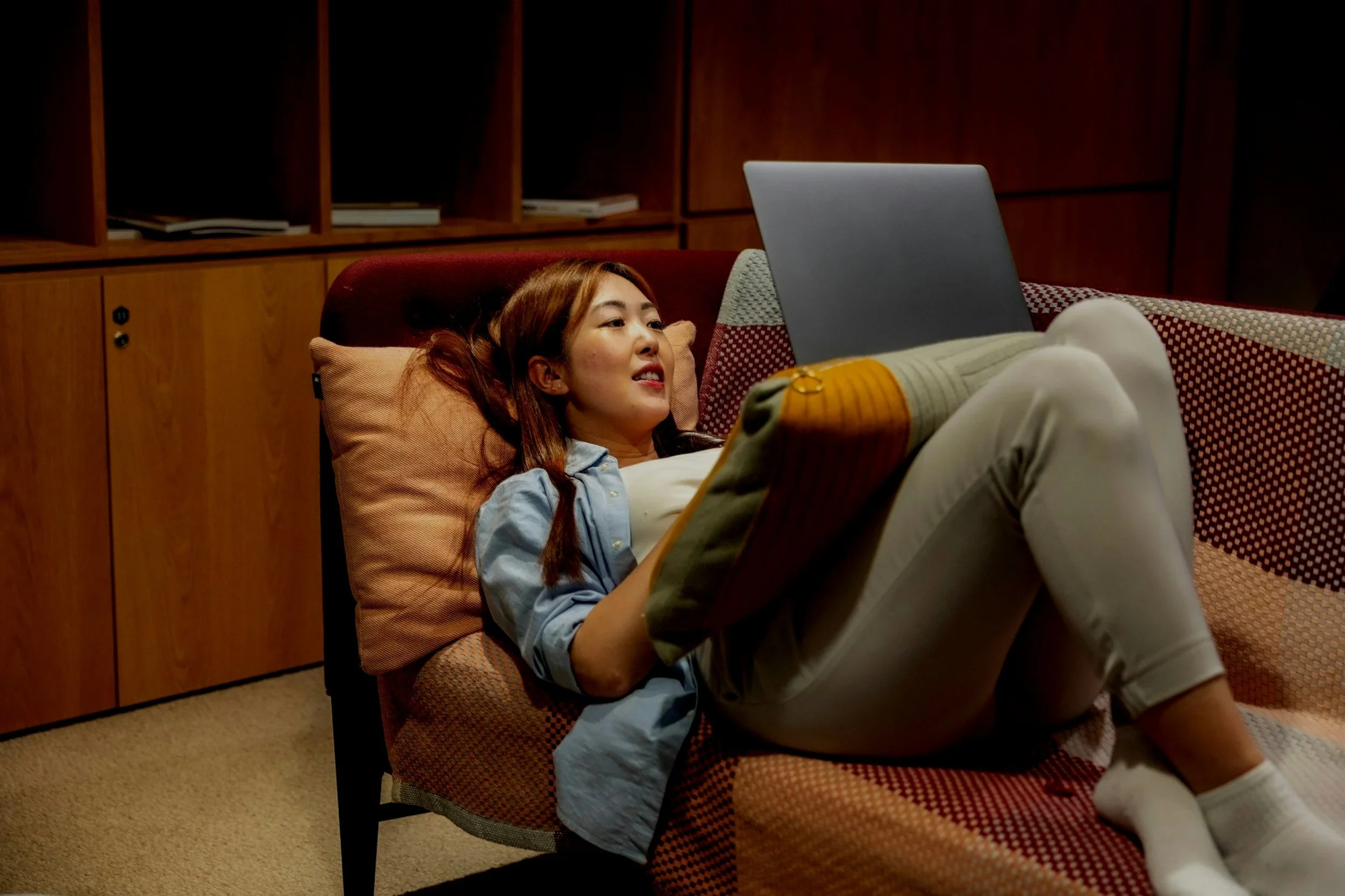 A young woman sitting cross-legged on a wooden platform in front of tall plants, using a silver Acer laptop.