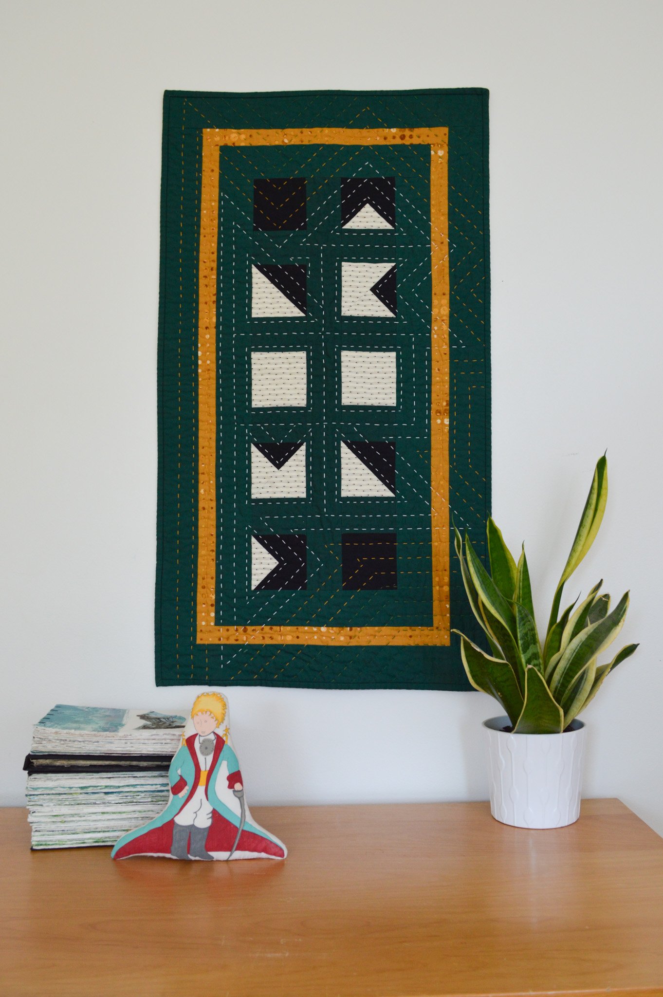  A green, quilted wall hanging with a snake plant, a stack of handmade books, and a Little Prince doll 