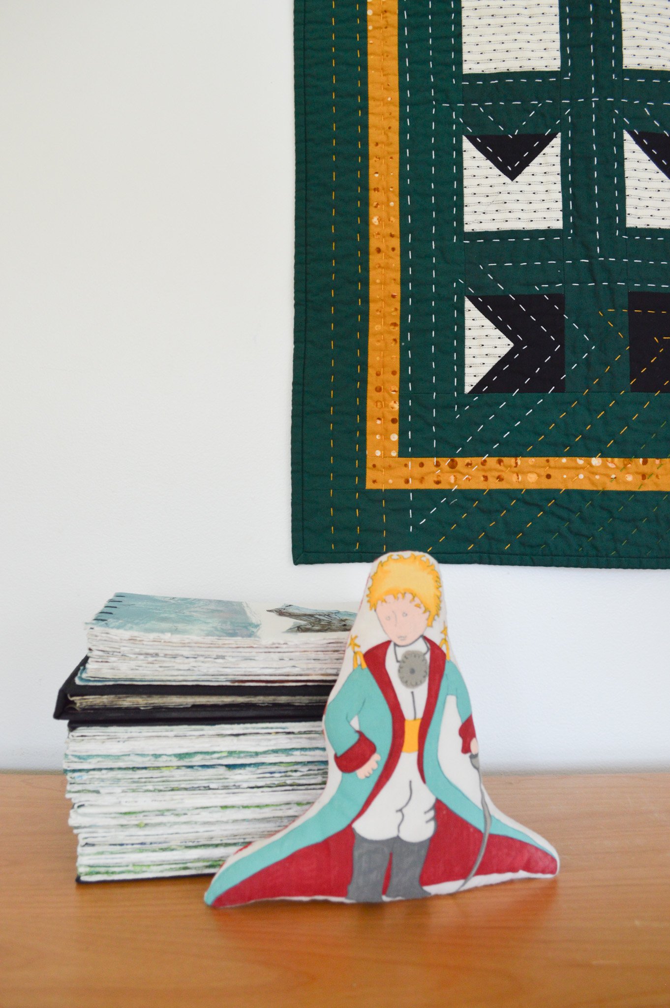  close up of a stack of handmade books and a Little Prince doll in front of a green, quilted wall hanging 