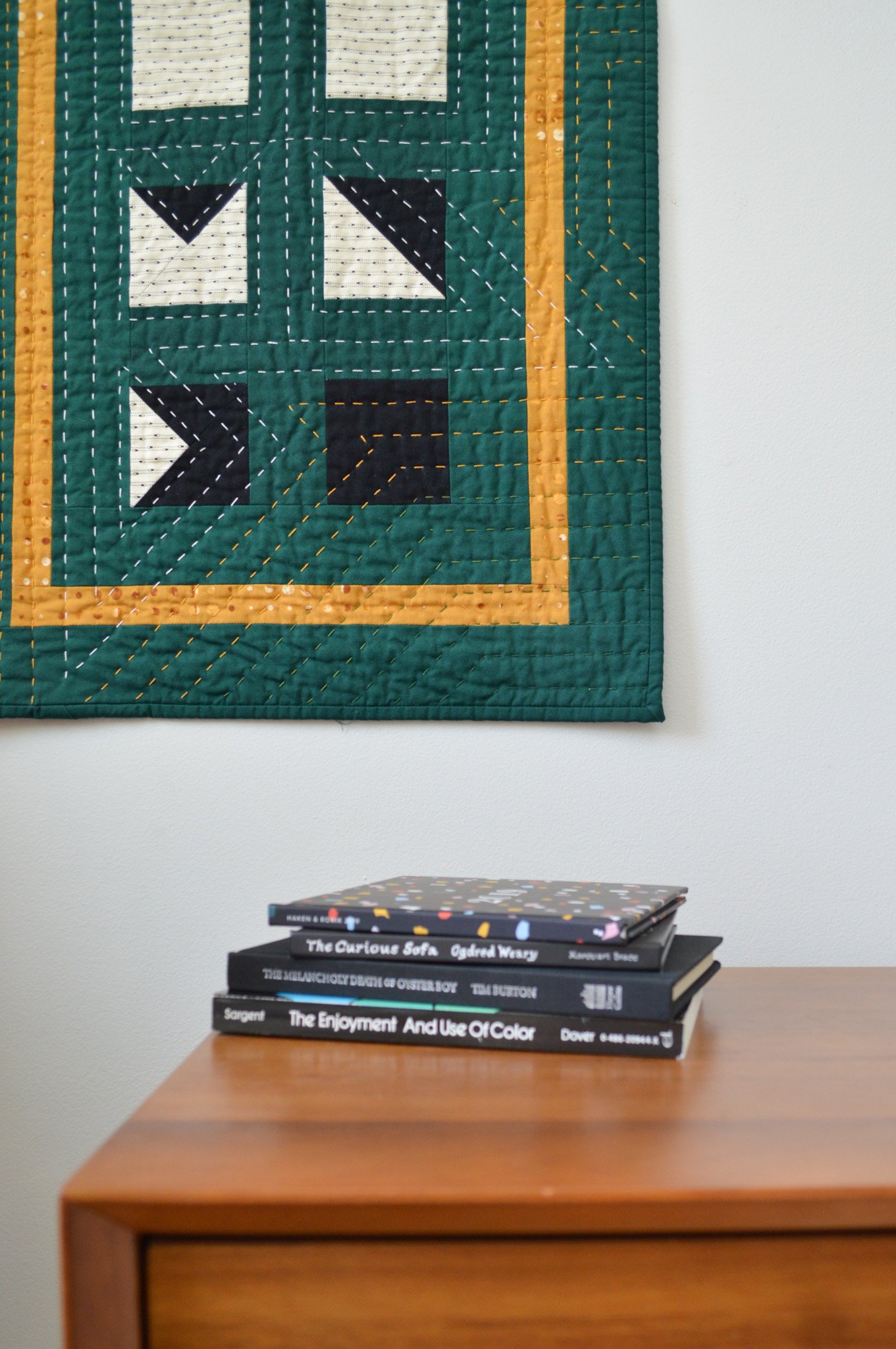  a stack of books on a modern side table with a green mini quilt wall hanging behind it 