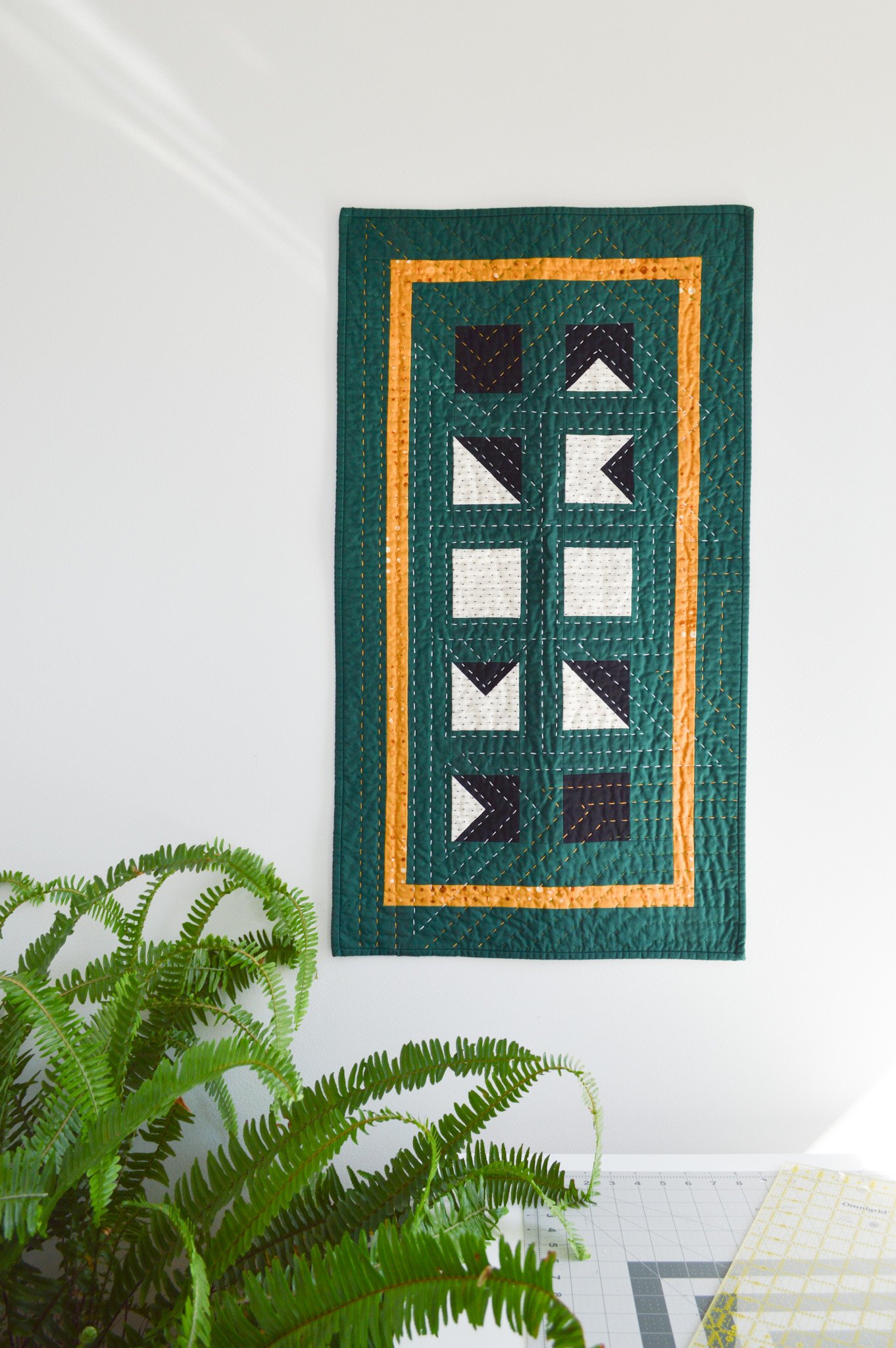  a green mini quilt hanging on a wall in a sewing room with a large fern 