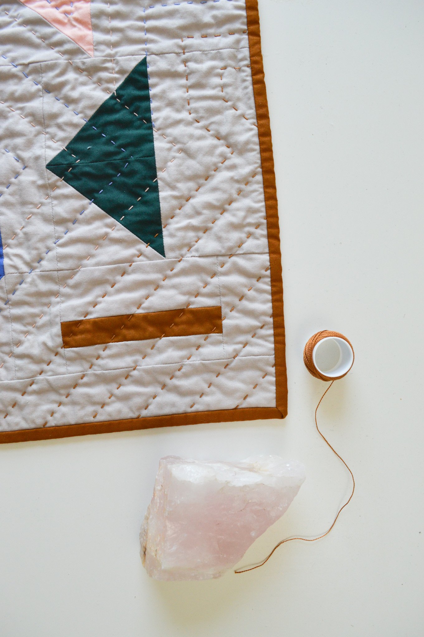  a hand quilted mini quilt on a white table with quilting thread and rose quartz 