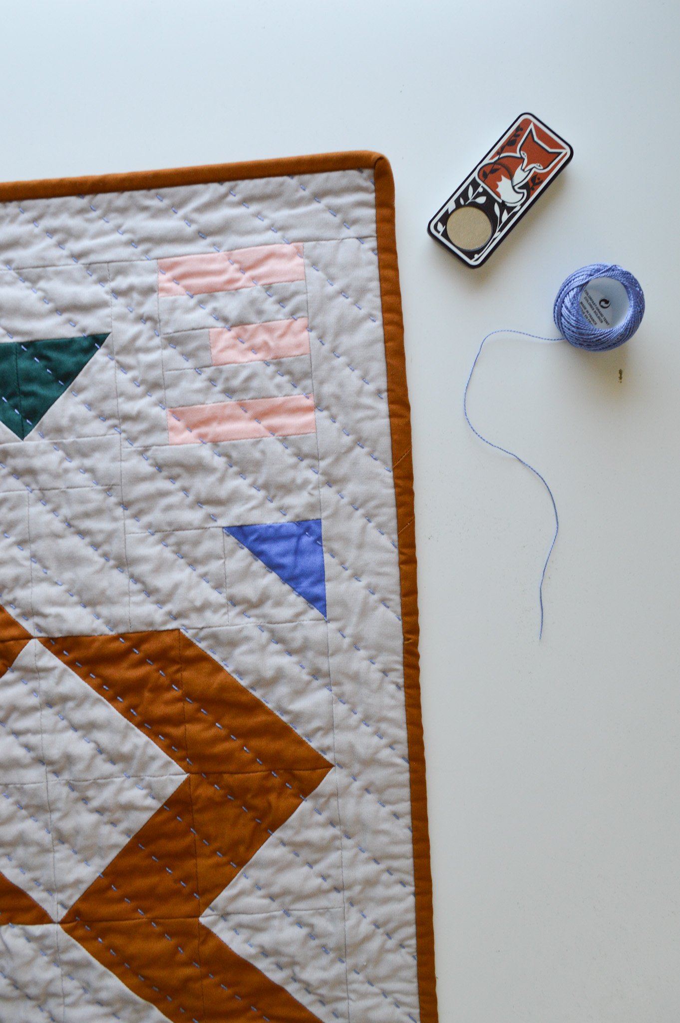  a hand quilted mini quilt on a white table with blue quilting thread and a needle box 