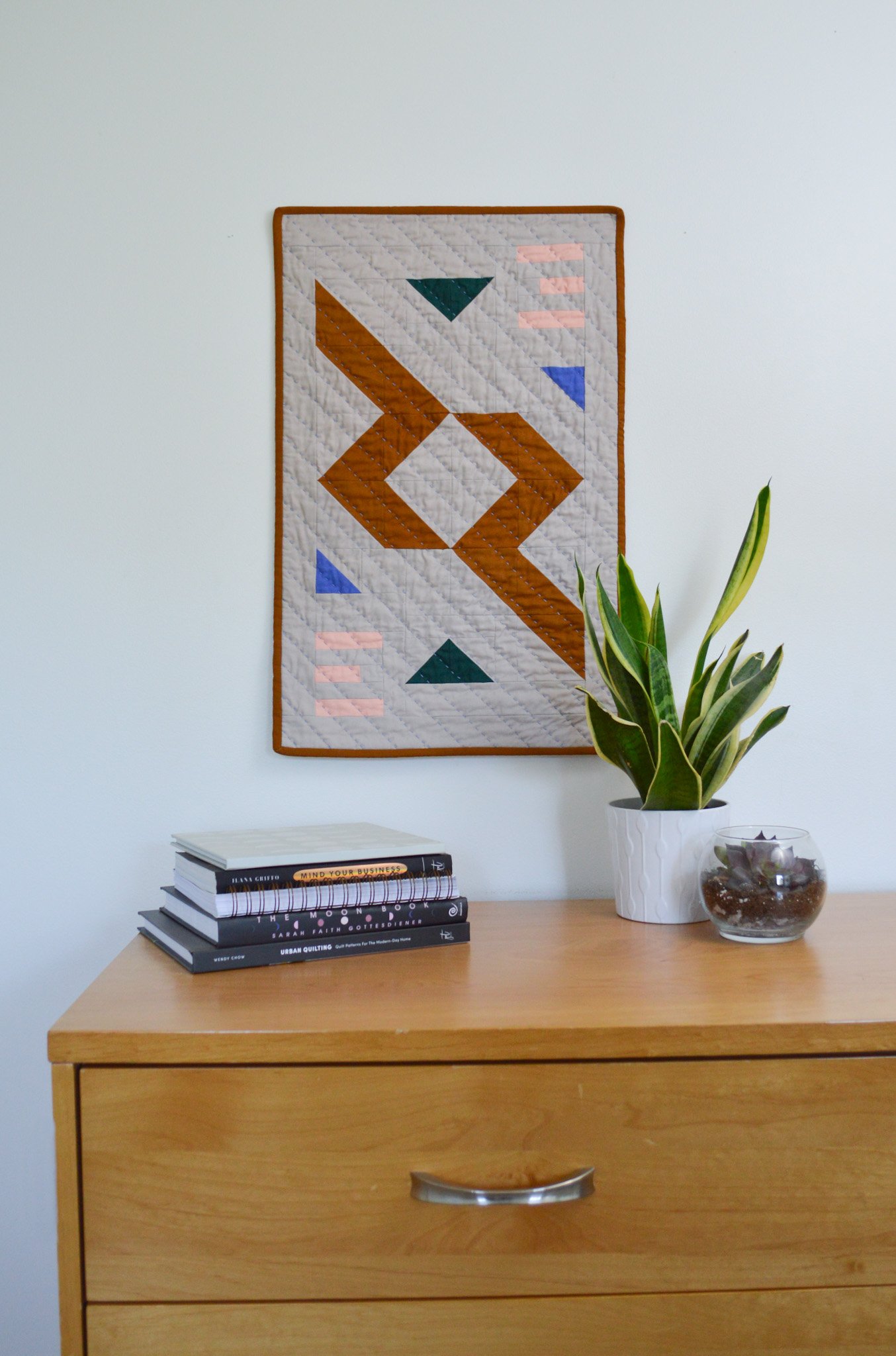  a modern, quilted wall hanging behind some plants and a stack of books 