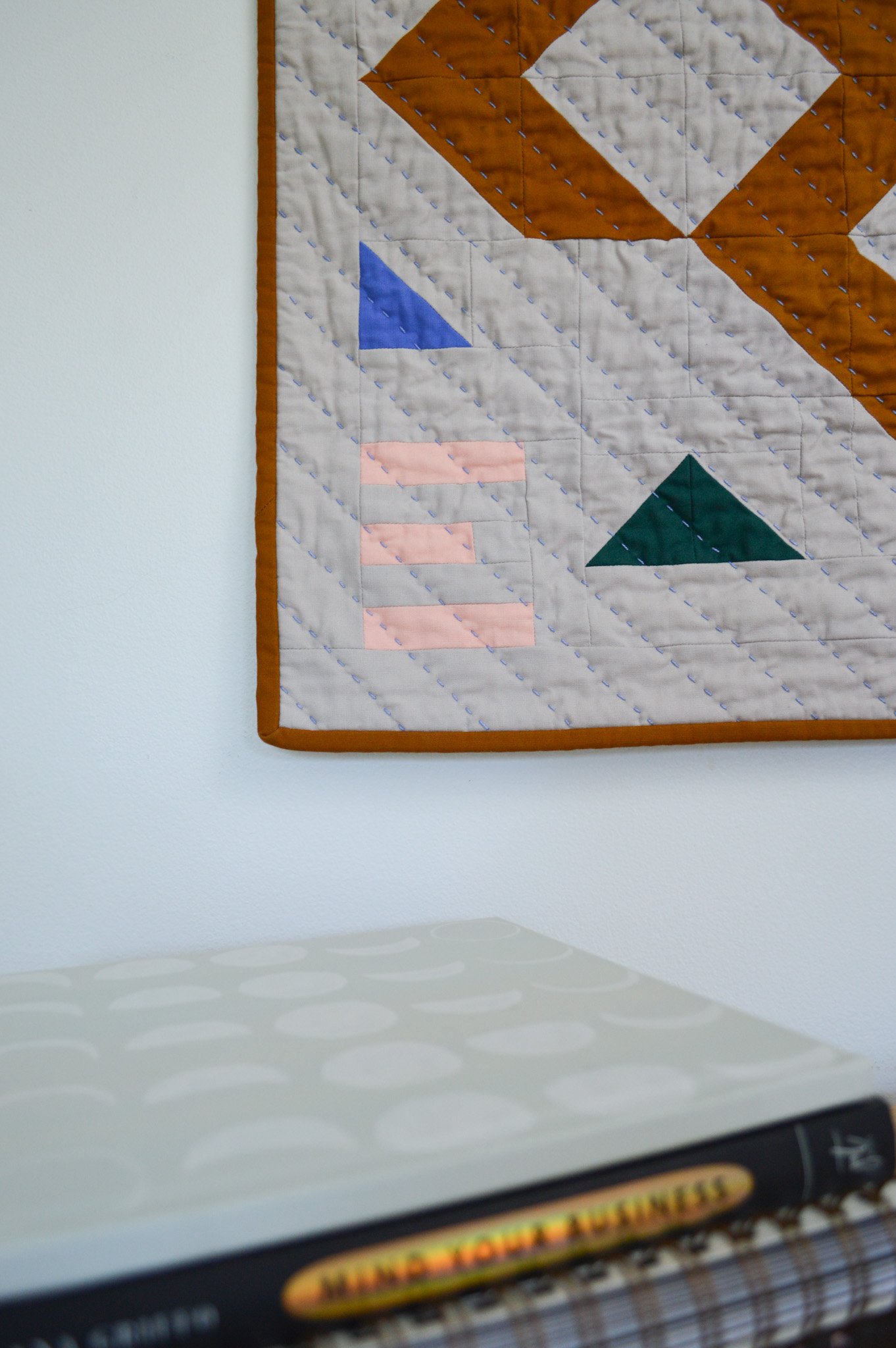  detail of hand stitching on a modern mini quilt handing behind a stack of books on a dresser 