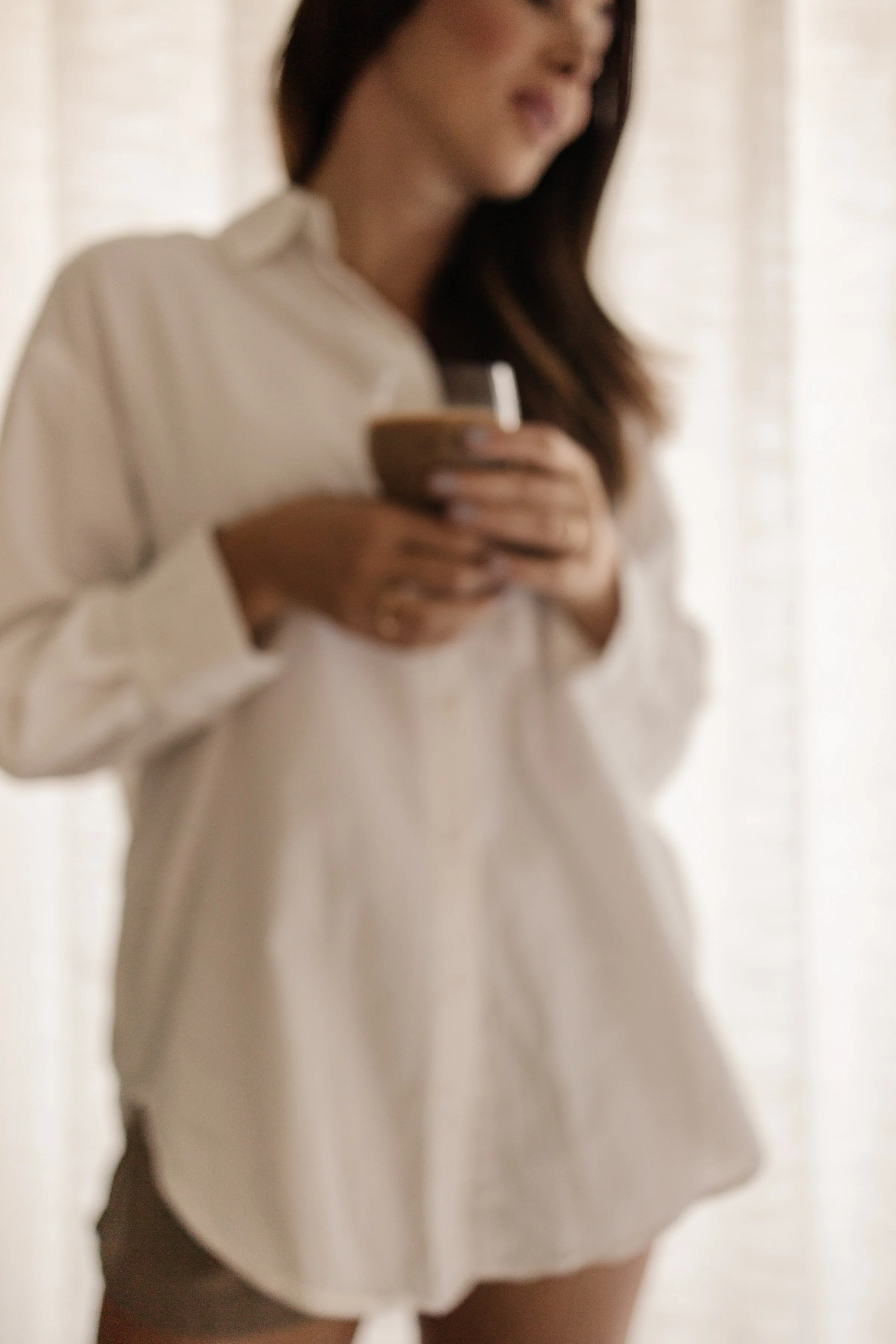A woman in a white shirt holding a cup, standing in front of a light-colored curtain.