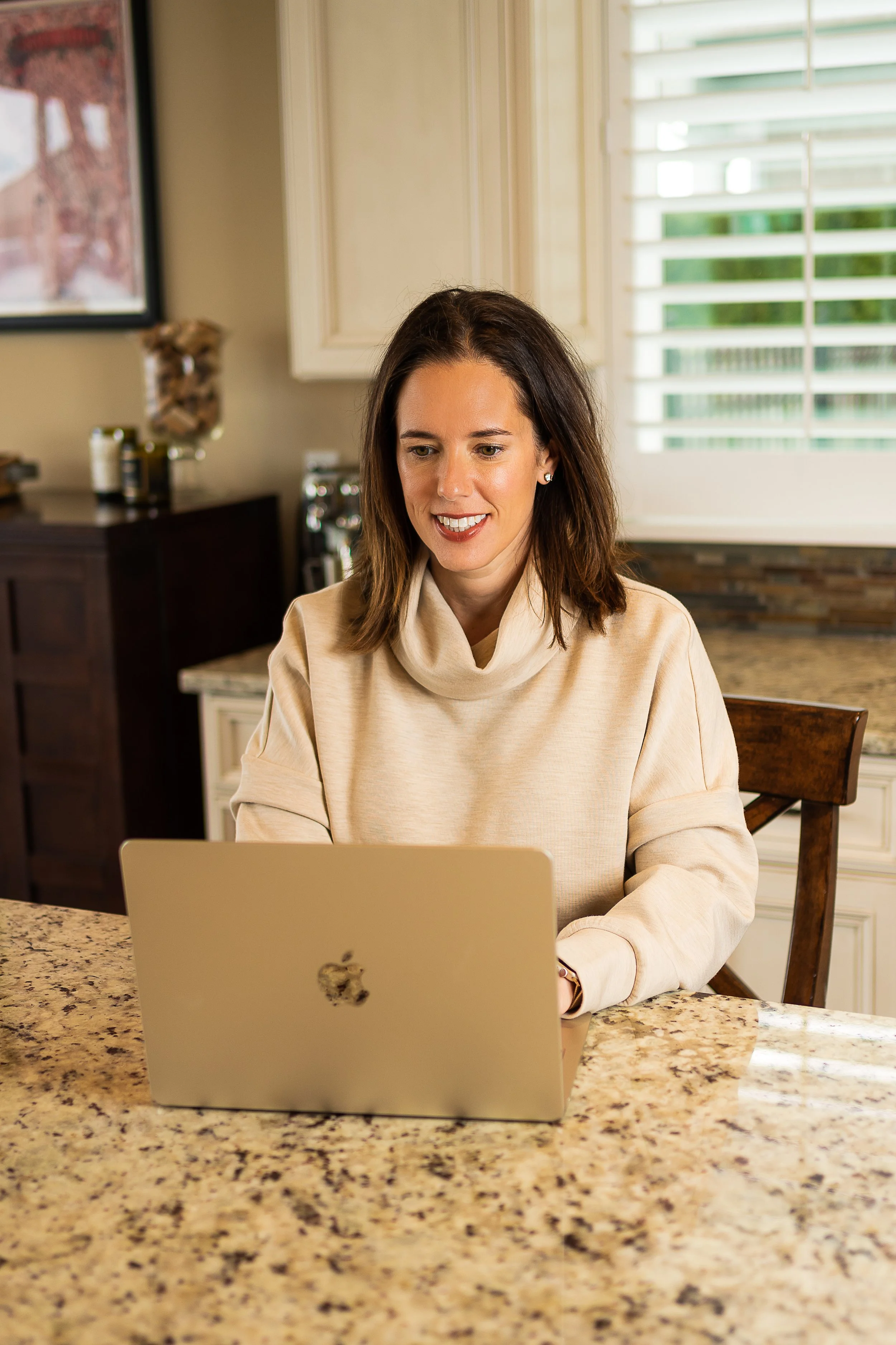 A woman sitting at a kitchen counter using a silver MacBook laptop, smiling, with a window with white shutters in the background.