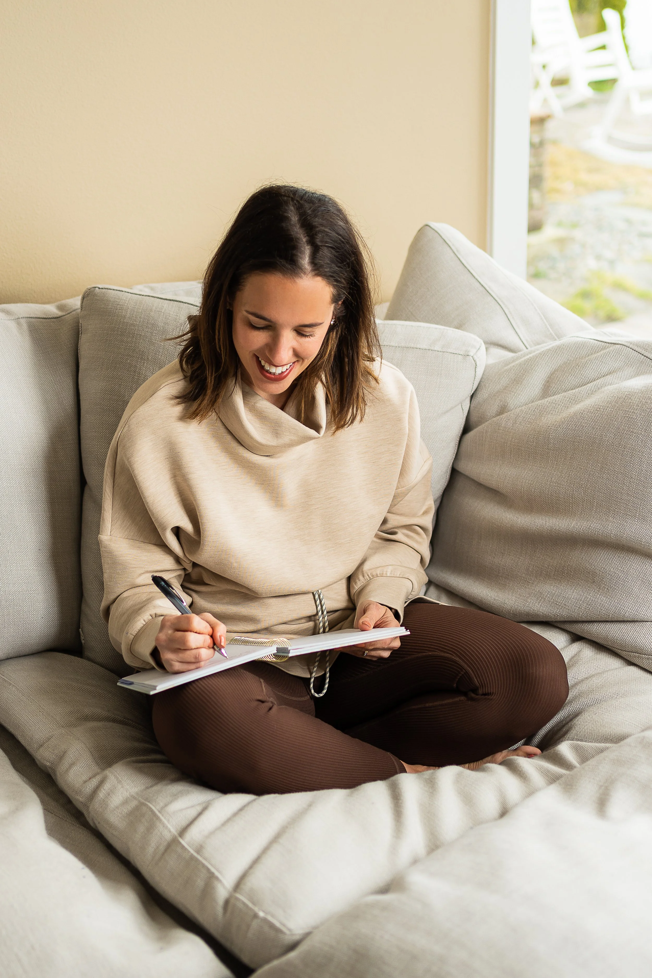 A woman with shoulder-length brown hair sitting on a light-colored couch, smiling while writing in a notebook with a pen, in a cozy indoor setting near a window.