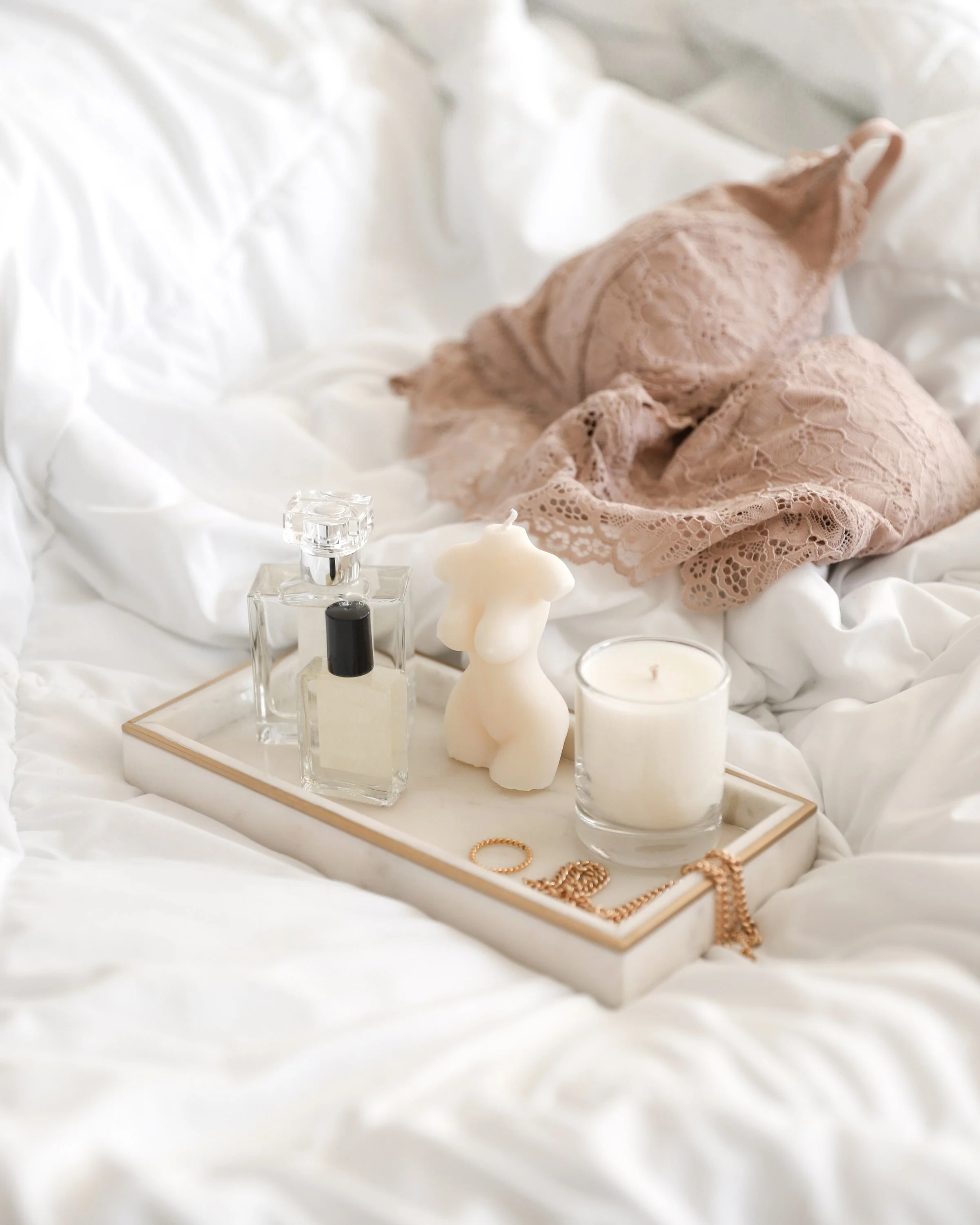 A bedroom scene with a white tray holding perfume bottles, a lit candle, a small torso-shaped candle, a gold ring, and a gold chain, set on white bedding with pink lace lingerie in the background.