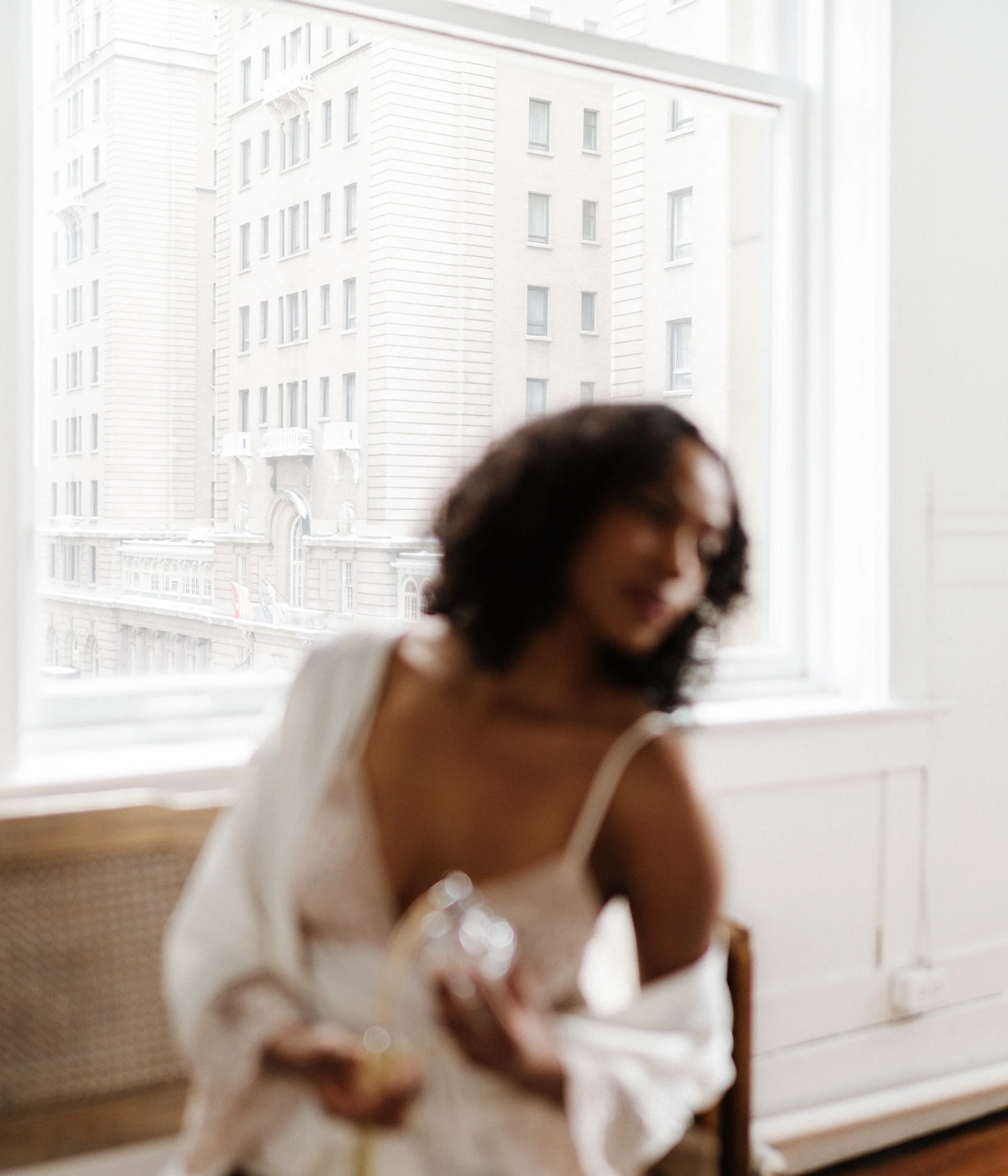 A woman with dark curly hair wearing a white dress sitting by a window with city buildings outside.
