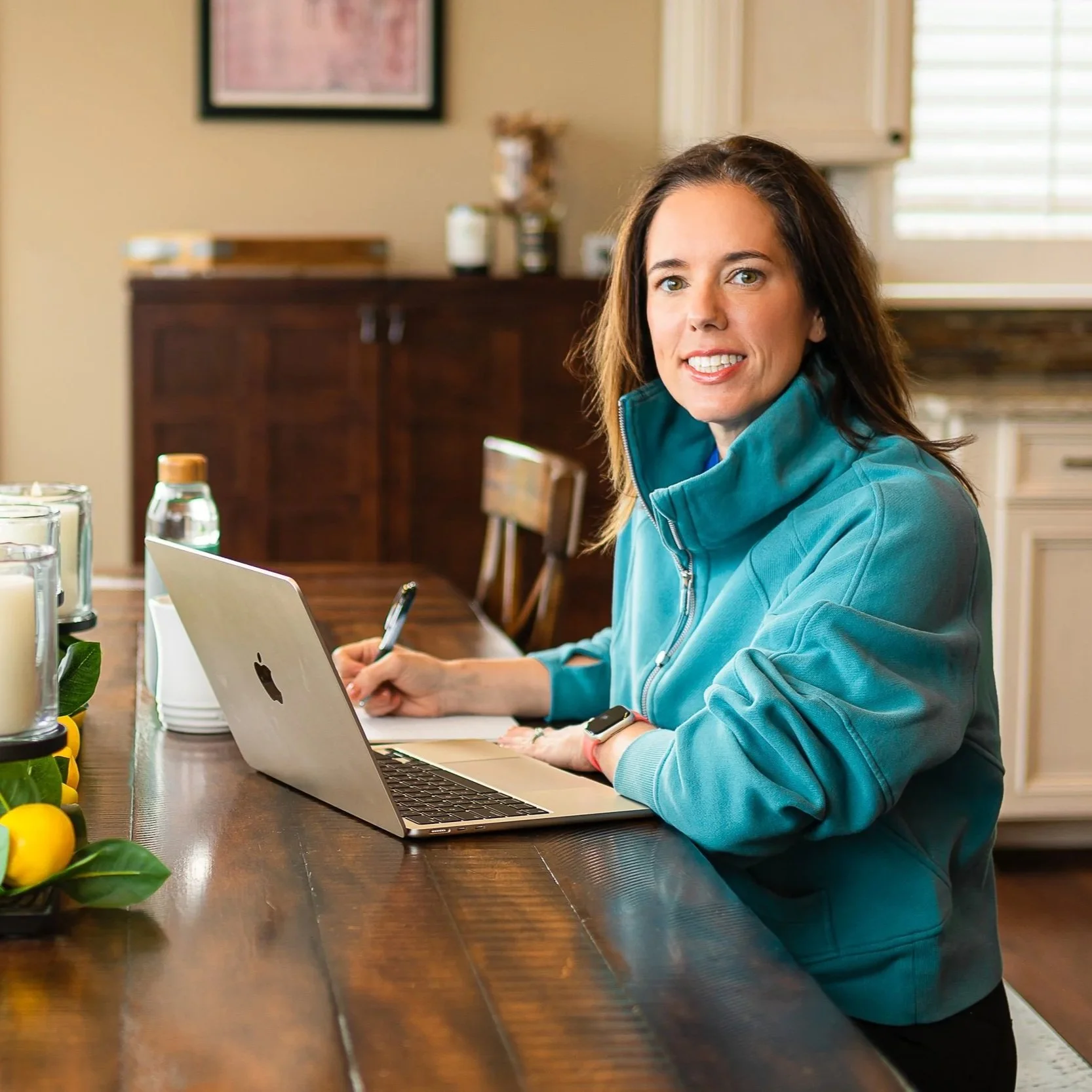 Woman in a blue zip-up jacket working at a wooden kitchen table with a MacBook and a pen, smiling at the camera.