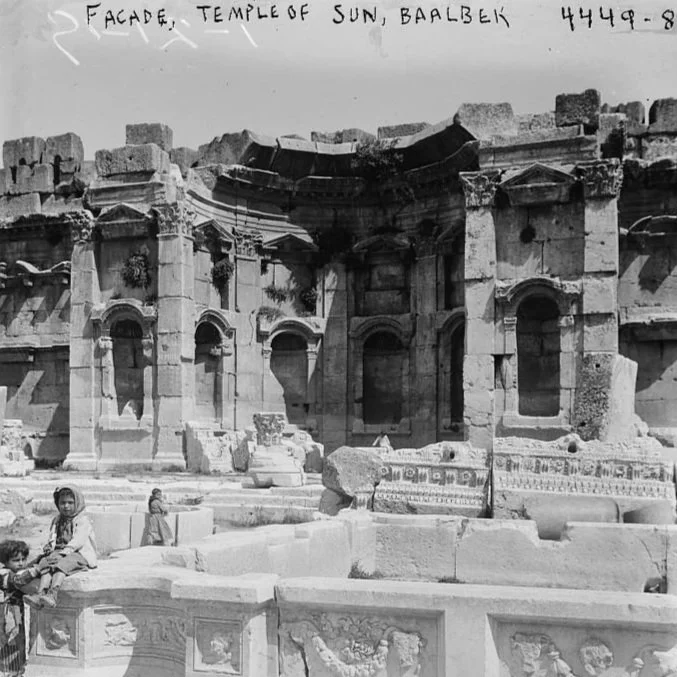 Black and white photograph of the ruins of the Temple of Baalbek, showing a large stone facade with arches and columns, and several children in traditional clothing sitting and standing in the foreground.