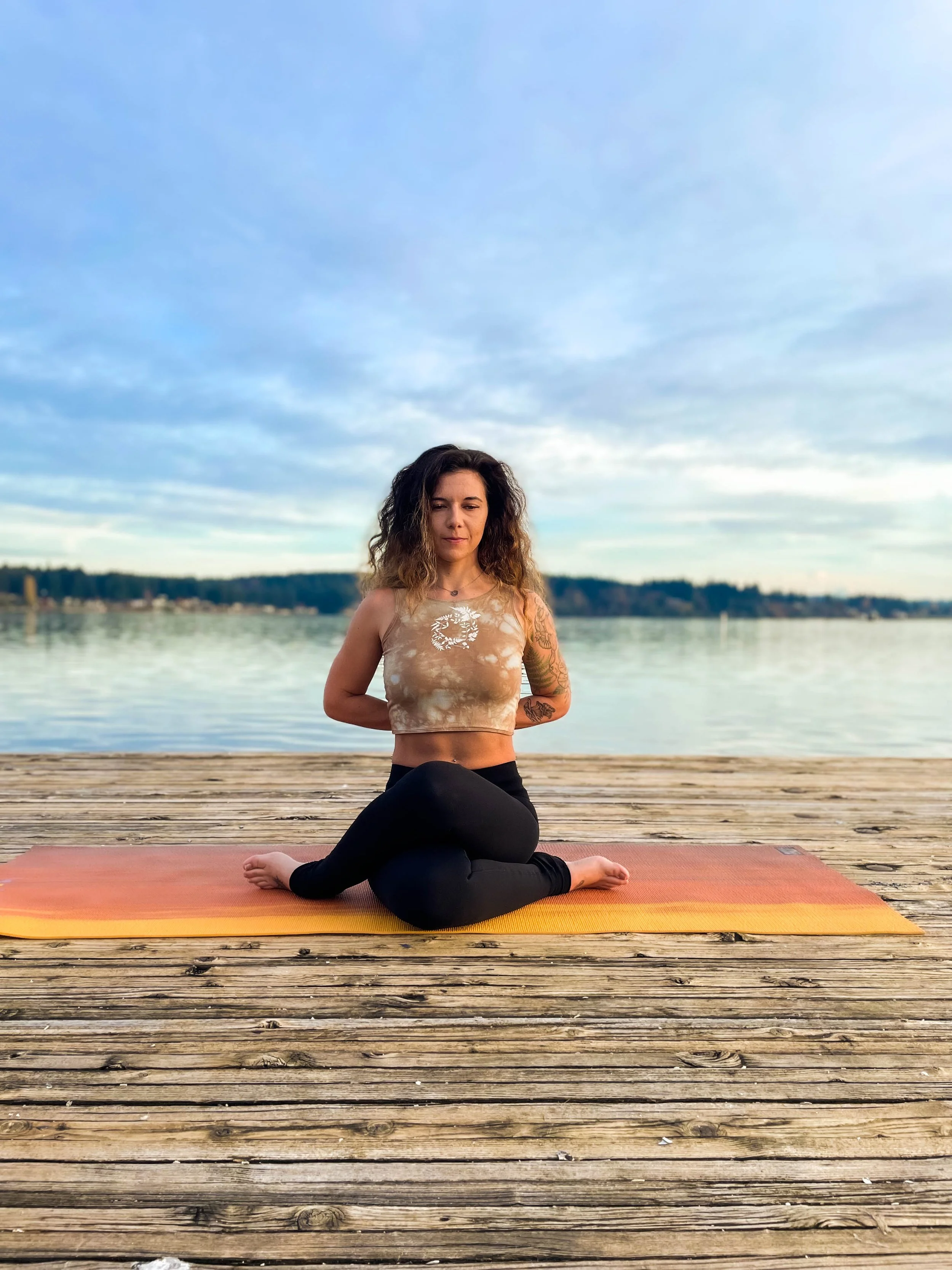 A woman appears to be performing a yoga pose on a wooden dock by the water with a background of trees and a cloudy sky.