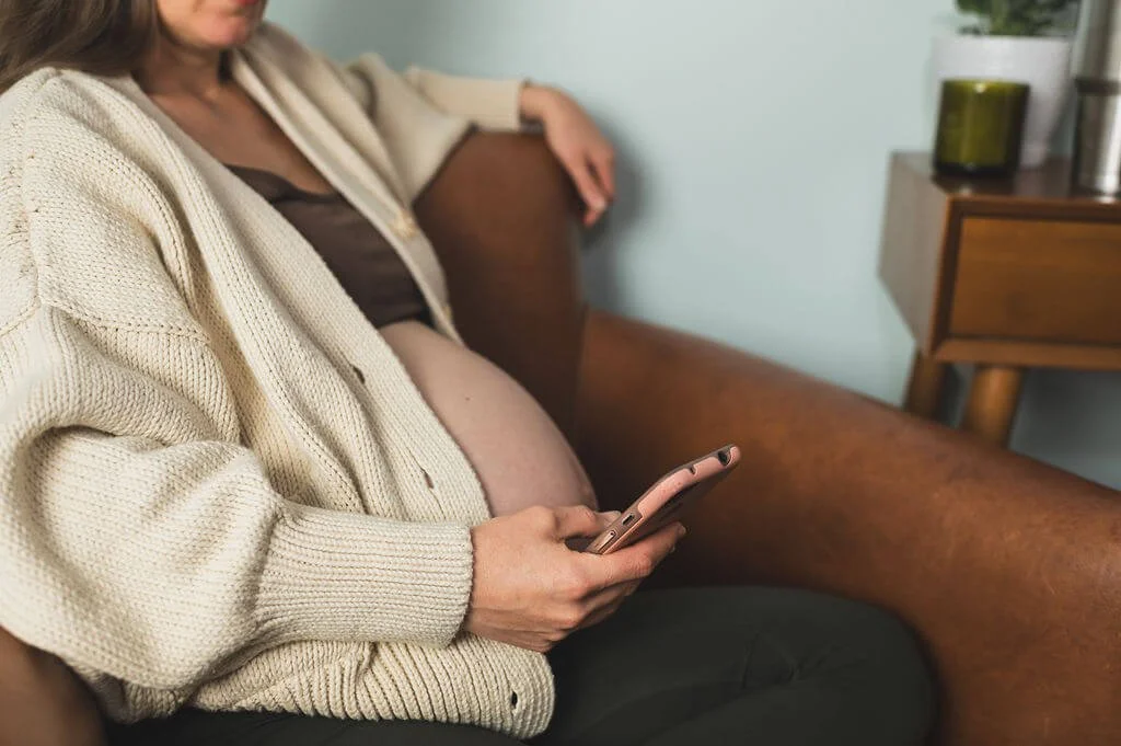 Pregnant woman wears tan sweater and reviews Progressive Pregnancy on her pink cell phone while she sits in a brown chair.
